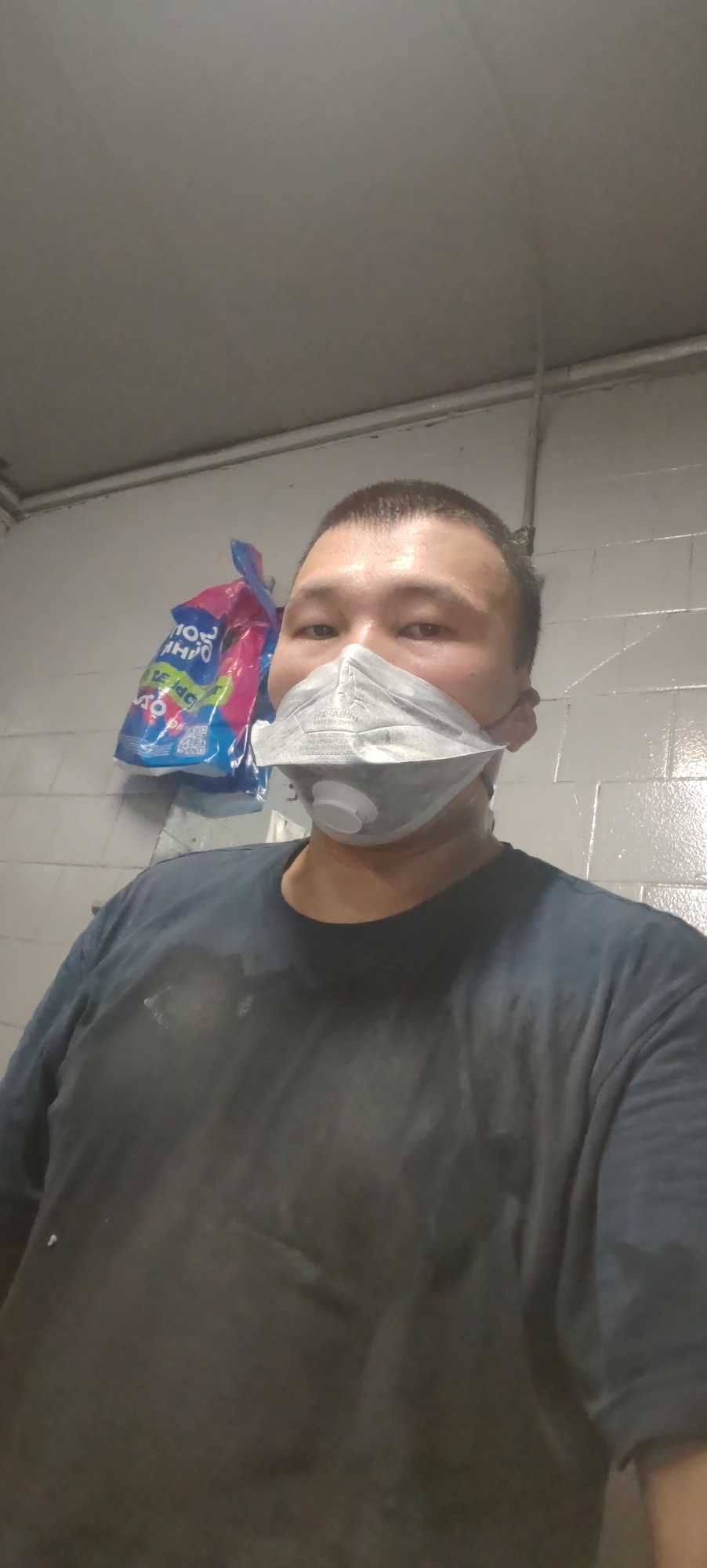 A man wears a grey respirator mask and a dark t-shirt. He stands in front of a tiled wall, with a large, brightly colored bag hanging in the background. The man's t-shirt appears to be dusty or dirty, with visible lighter patches. The lighting suggests an indoor environment, possibly a workshop or storage area. No specific location in Поселье, Russia, can be identified from the visual evidence.