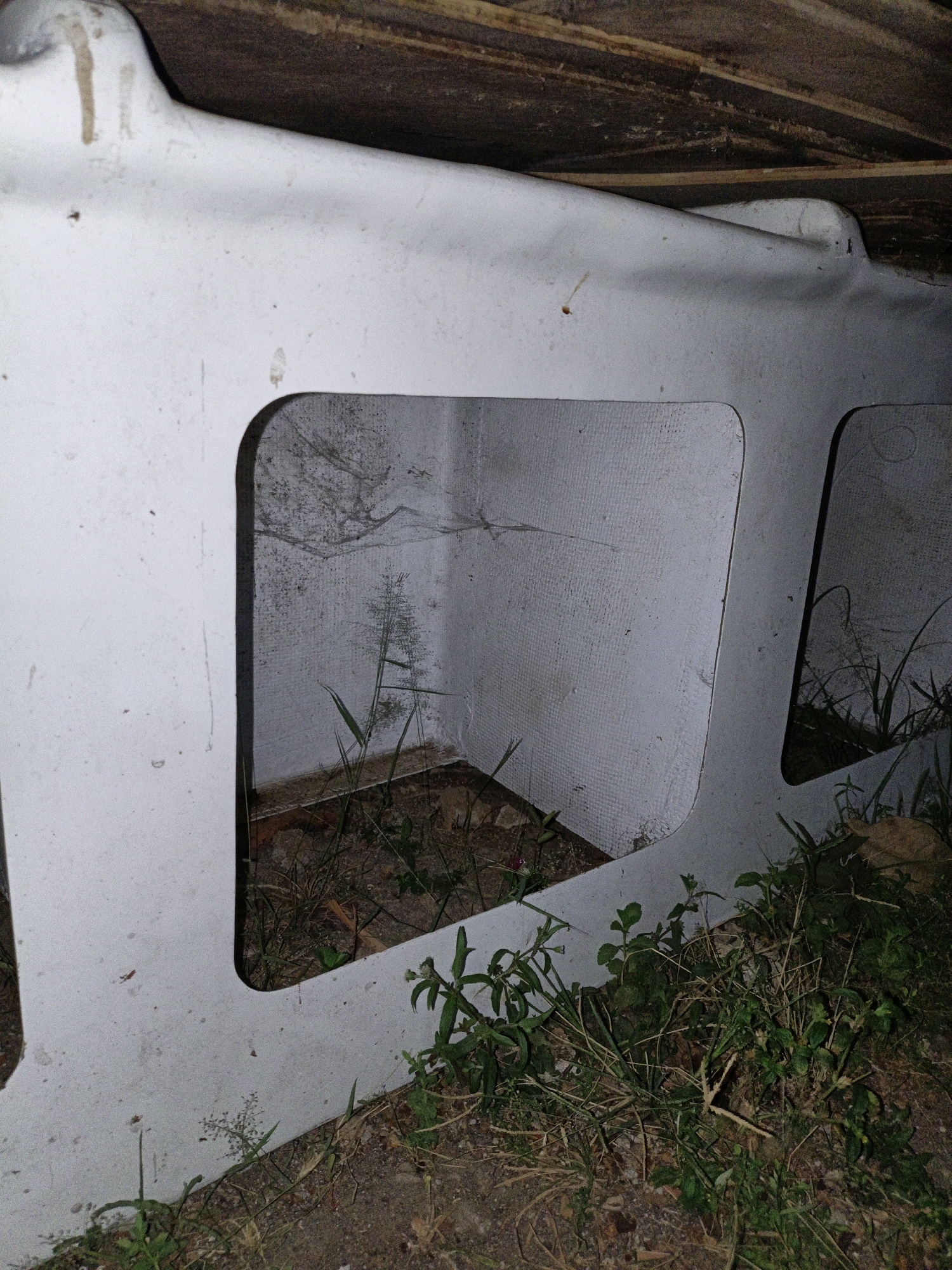 The image shows a side view of a large, white, possibly fiberglass, structure with two recessed openings. The structure is supported by wooden beams at the top, suggesting it is part of an overhead construction or shelter. The ground in front and around the structure is covered in dirt, small rocks, and overgrown vegetation including grasses and small leafy plants. Through the left opening, the interior of a small, square, white-walled space is visible. The walls of this interior space appear rough and have some cobwebs and faint markings. A thin, feathery grass stalk is growing inside this opening. The right opening reveals a similar interior space, also with vegetation growing in front of it and extending slightly into the visible area. The lighting suggests it is either dusk or a dimly lit enclosed space. The scene appears to be outdoors in an unkempt area, possibly an abandoned or disused part of an industrial or construction site. The presence of overgrown vegetation and the general state of disrepair suggest this location is not regularly maintained. It is not possible to determine the specific location within Cikal, Indonesia from the image alone.