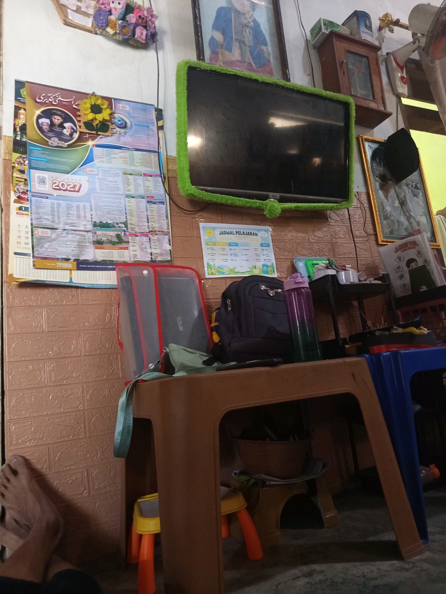 The image depicts an indoor scene, likely a room in Ngablak, Indonesia. In the upper left corner, a wall-mounted display features colorful felt flowers arranged as an owl and other decorative elements. To the right of this display, a framed picture with a blue-clad figure is visible. Dominating the upper portion of the image is a television screen with a bright green fuzzy border, mounted on the wall. Below the television, a framed picture with a gold border shows a person in a white robe.
A prominent feature in the center of the image is a calendar with the year "2027" clearly visible. The calendar is adorned with a yellow sunflower decoration. Next to the calendar, a blue and white poster titled "JADWAL PELAJARAN" (meaning "Lesson Schedule") is displayed.
In the foreground, a brown plastic table holds various items. A black backpack, a purple and green gradient water bottle, and a red and gray laptop sleeve are placed on the table. There is also a small green pouch and what appears to be a roll of tape. To the right of the table, a blue plastic chair is visible, with a small orange and yellow stool placed underneath the brown table. The floor appears to be a grey tiled surface.
On the far left of the image, a person's bare feet and lower legs are visible, suggesting someone is seated or lying down in that area. The walls are covered with a textured brown material, resembling brick or wood paneling. The overall lighting is dim, creating shadows throughout the scene.