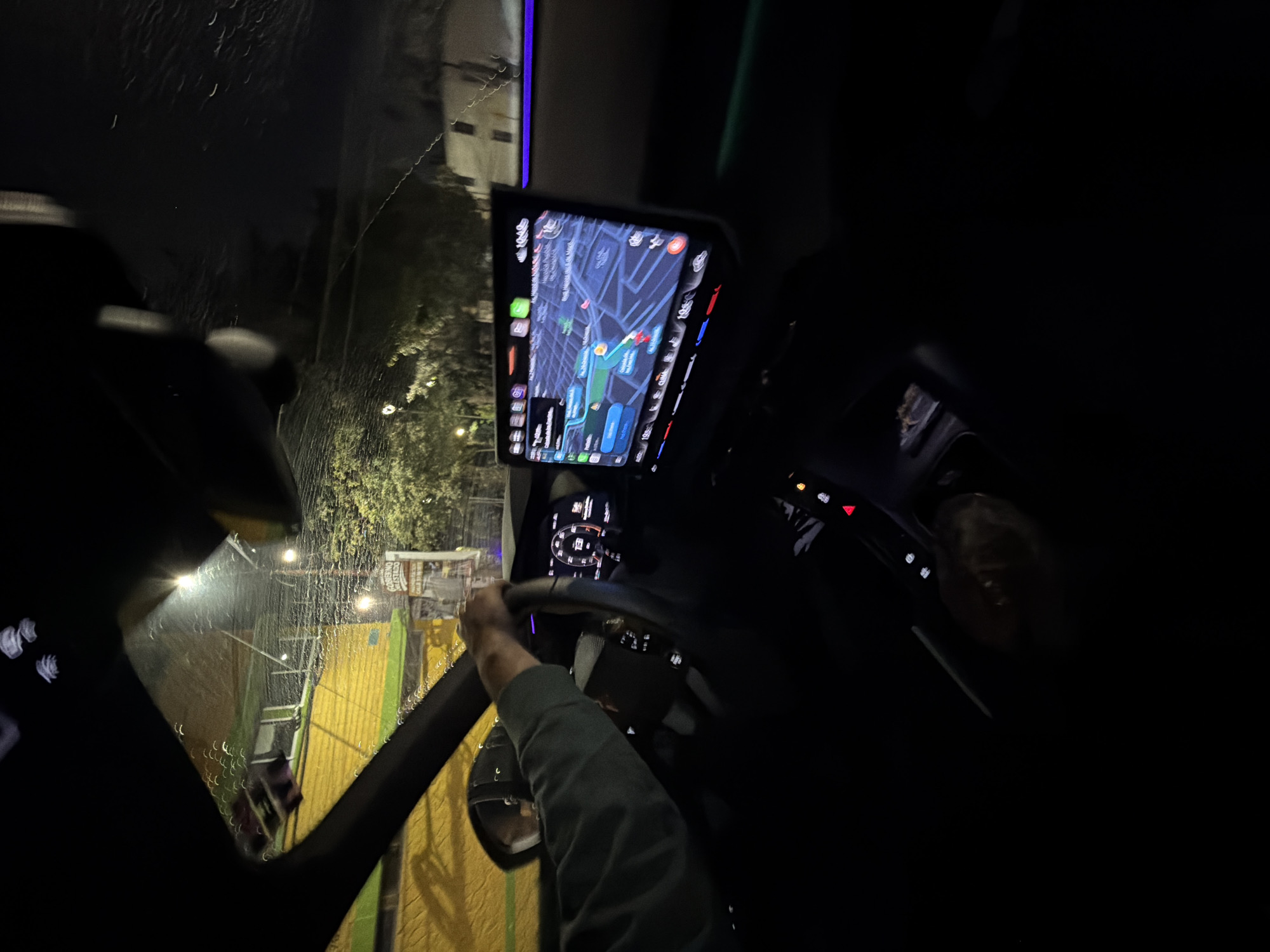 The image, taken at night, appears to be from inside a vehicle in Puebla, Mexico. Rain streaks the windshield, obscuring the view of a brightly lit outdoor area with yellow and green structures. A navigation screen in the vehicle displays a map of Puebla, indicating the vehicle's location and route. The driver's hands are visible on the steering wheel, with one arm clad in a long-sleeved olive green shirt. The vehicle's dashboard and interior elements are also visible, but largely obscured by darkness.