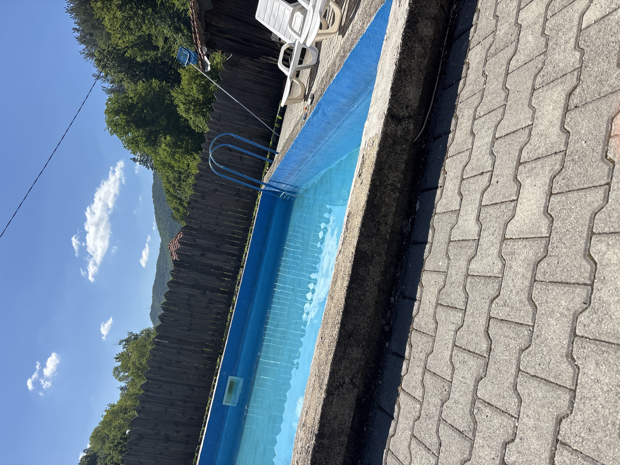 The image shows an outdoor swimming pool located in Liava Reka, Bulgaria. The pool is rectangular with bright blue lining and filled with clear blue water. A metal ladder with blue steps is attached to the side of the pool. The pool area is paved with gray interlocking concrete pavers. To the right of the pool, two white plastic lounge chairs are visible. Behind the pool, there is a wooden fence and a dense green forest covering rolling hills under a bright blue sky with scattered white clouds. A portion of a rustic wooden structure with a brown roof is also visible to the right, partially obscured by trees. A blue pool skimmer on a pole is propped against this structure.