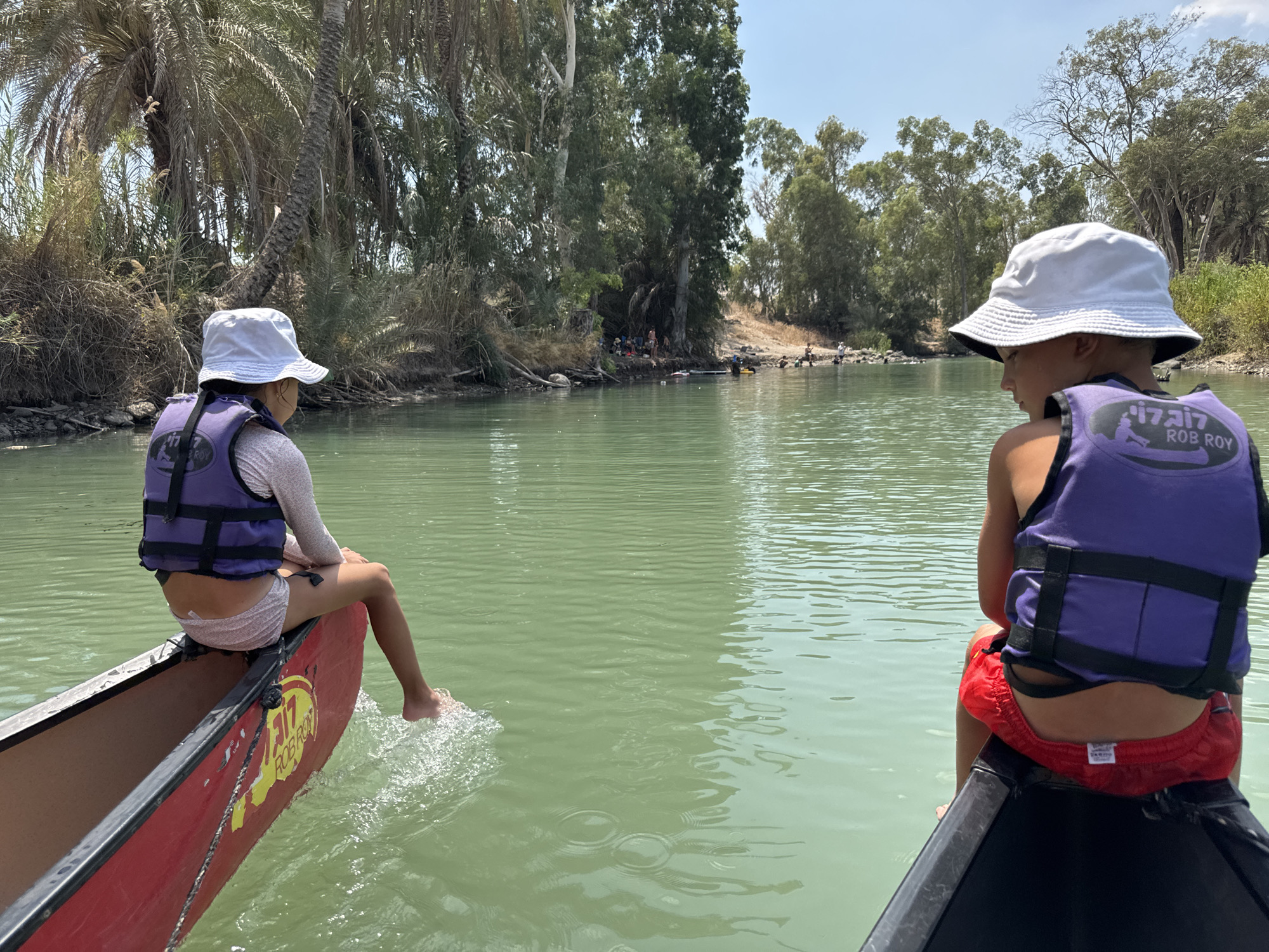 In the city of Emek HaYarden Regional Council, Israel, two children, one with their back to the camera and the other facing away, are seated at the front of two canoes on a calm, green body of water. Both children are wearing white bucket hats and purple life vests. The child on the left is wearing light-colored shorts and has their feet trailing in the water, creating ripples. The child on the right is wearing red swimming trunks. The canoes appear to be moving. The shoreline in the background is lined with lush green trees, including palm trees, and dense vegetation. Several other people are visible on the shore in the distance, some appearing to be interacting with the water. The sky is clear and blue, suggesting a sunny day.