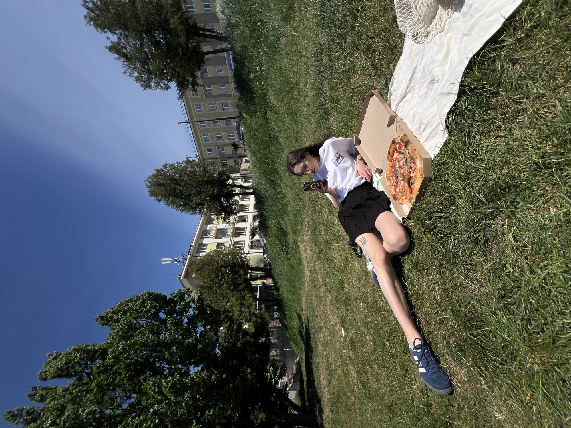 A woman is sitting on a grassy hillside in Ternopil, Ukraine, with a pizza box resting next to her on a white cloth. The woman is wearing a white t-shirt, black shorts, and blue athletic shoes. She is holding a smartphone and appears to be looking at it. Beside her is an open pizza box showing a pizza. In the background, apartment buildings are visible. The sky is clear and blue.