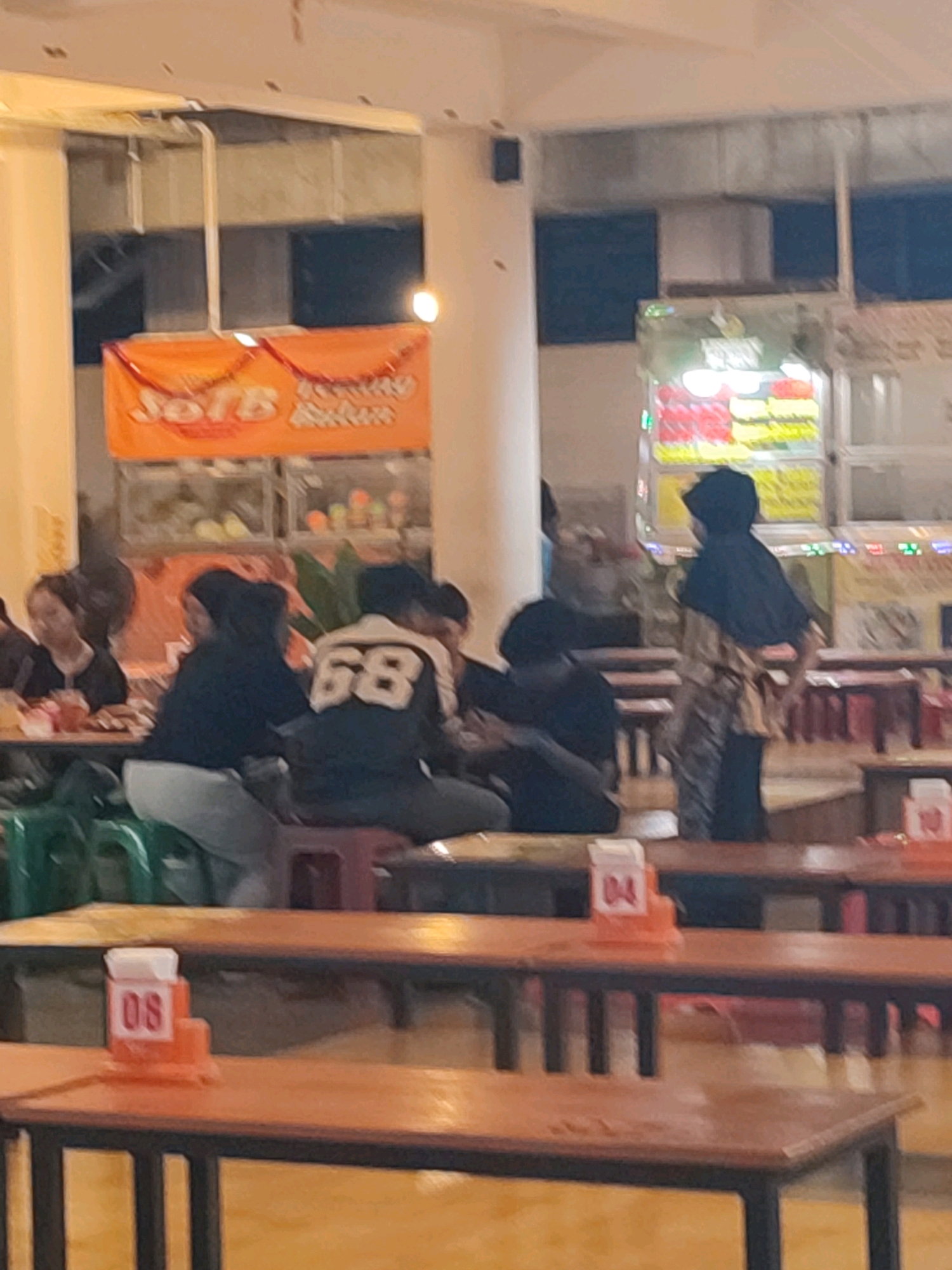 The image depicts an indoor setting, likely a food court or restaurant, with several people visible. In the foreground, a wooden table with black metal legs is prominently displayed, with an orange table number holder labeled "08" on it. To the right, another similar table is visible, with an orange table number holder labeled "04". Behind these tables, a group of people are seated. One person in the center is wearing a black jacket with the number "68" on the back. To their left, a person wearing a dark headscarf is seated, and further left, another person is partially visible. In the background, an orange banner with red lettering is hung, and a display case filled with various items, possibly food or drinks, is visible to the right. The overall lighting suggests an evening or indoor ambiance. The scene is consistent with a casual dining establishment in Yogyakarta, Indonesia, where such casual eateries are common.