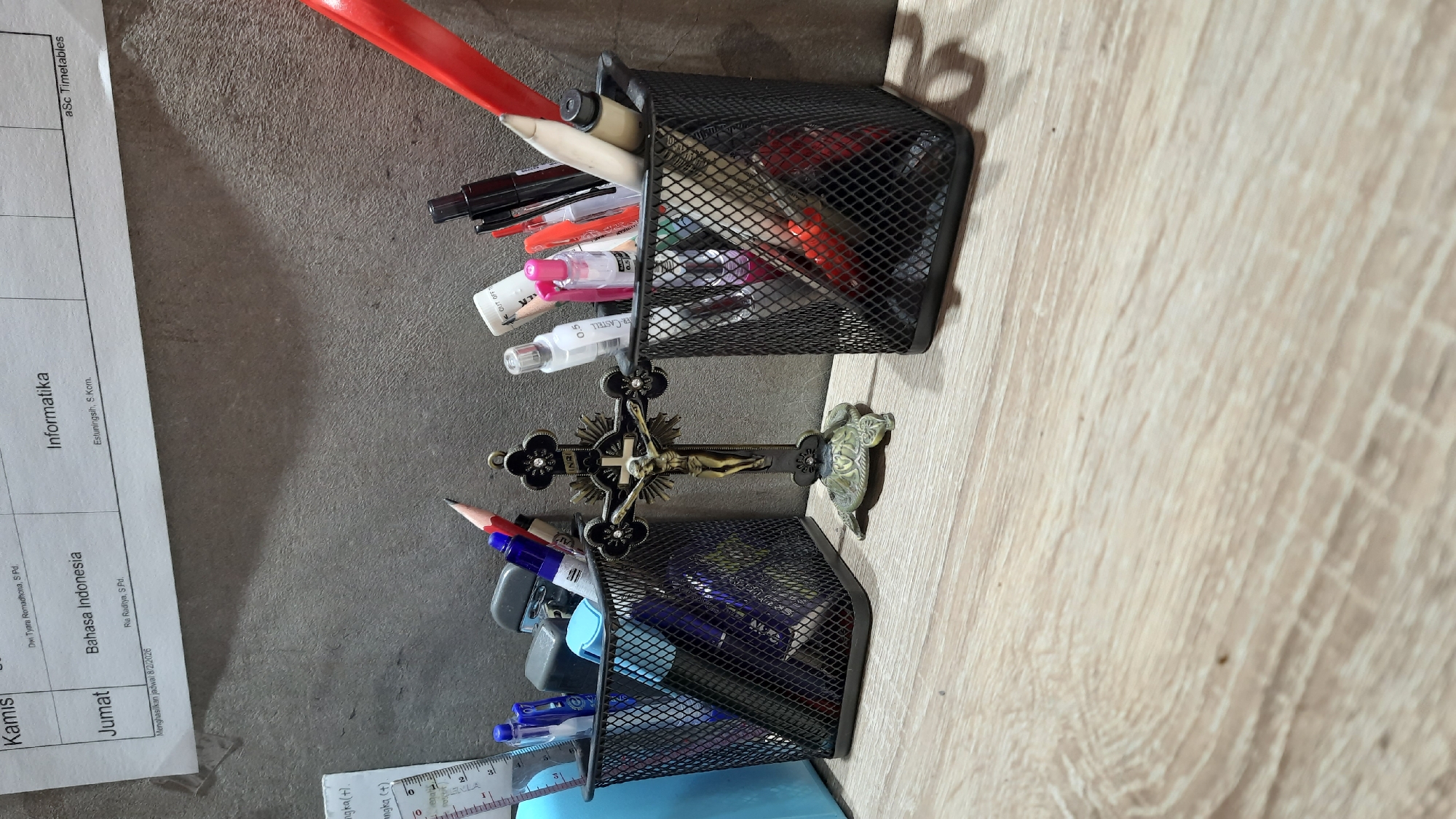 A close-up, eye-level shot captures a desk with two black mesh pen holders filled with various writing utensils. To the left, a blue container is partially visible. Between the pen holders stands a small, ornate crucifix with a figure of Jesus. A small bronze turtle figurine sits at the base of the crucifix. The pen holders and crucifix are positioned on a wooden desk surface. In the background, a textured gray wall is visible, with a portion of a paper schedule or timetable tacked to it at the top. The schedule lists subjects like "Kamis," "Dwi Tyara Romadhona, S.Pd," "Informatika," "Jumat," "Bahasa Indonesia," "Ria Raditya, S.Pd," and "aSc Timetables." Text indicating "Menghasilkan jadwal 8/2/2026" is also present on the schedule. The lighting suggests an indoor environment, possibly a home office or classroom.