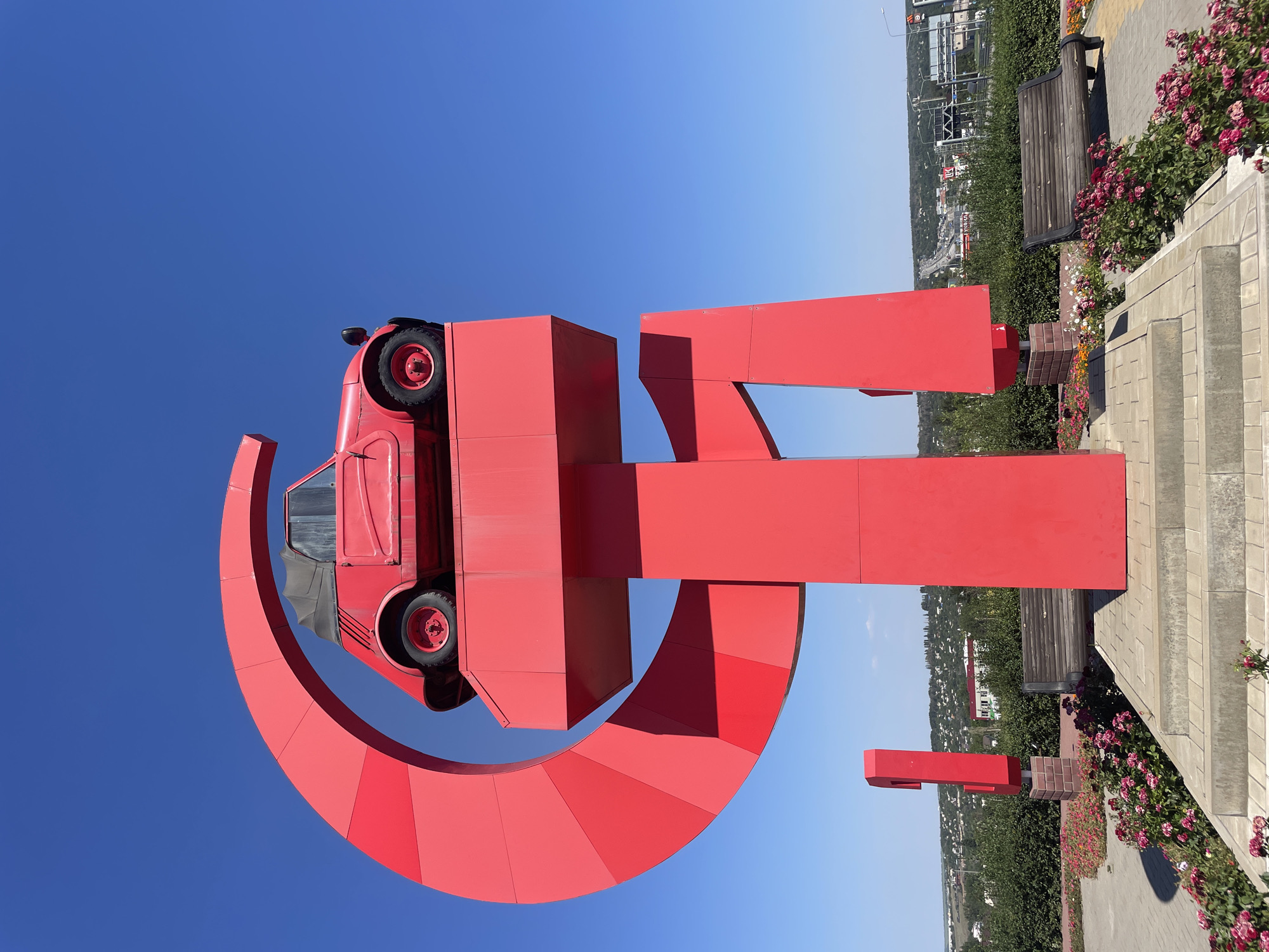The photograph captures the "KamAZ" monument in Kamensk-Shakhtinsky, Russia. The monument features a bright red, vintage KamAZ car perched atop a large, red, abstract sculpture. The sculpture is composed of geometric shapes, including a large crescent-like form and a vertical pillar, all painted in a bold red. The car itself is a small, two-door convertible with its top down. The monument stands against a clear, vibrant blue sky on a sunny day.
In the foreground, a paved walkway leads up to the monument, bordered by meticulously maintained flower beds bursting with pink and red roses. Two weathered wooden benches are placed along the walkway, inviting viewers to rest. To the left of the main monument, another, smaller red sculptural element resembling the numeral "1" stands on a brick base.
In the background, a wide view of Kamensk-Shakhtinsky unfolds. Green trees and bushes form a dense line separating the monument area from the urban landscape. Beyond the greenery, houses with red roofs and other buildings of the city are visible, extending towards a distant highway with overhead signs. The overall impression is one of a well-maintained public space, a roadside attraction likely intended to draw attention and commemorate local industry or history. There are no people visible in the frame.