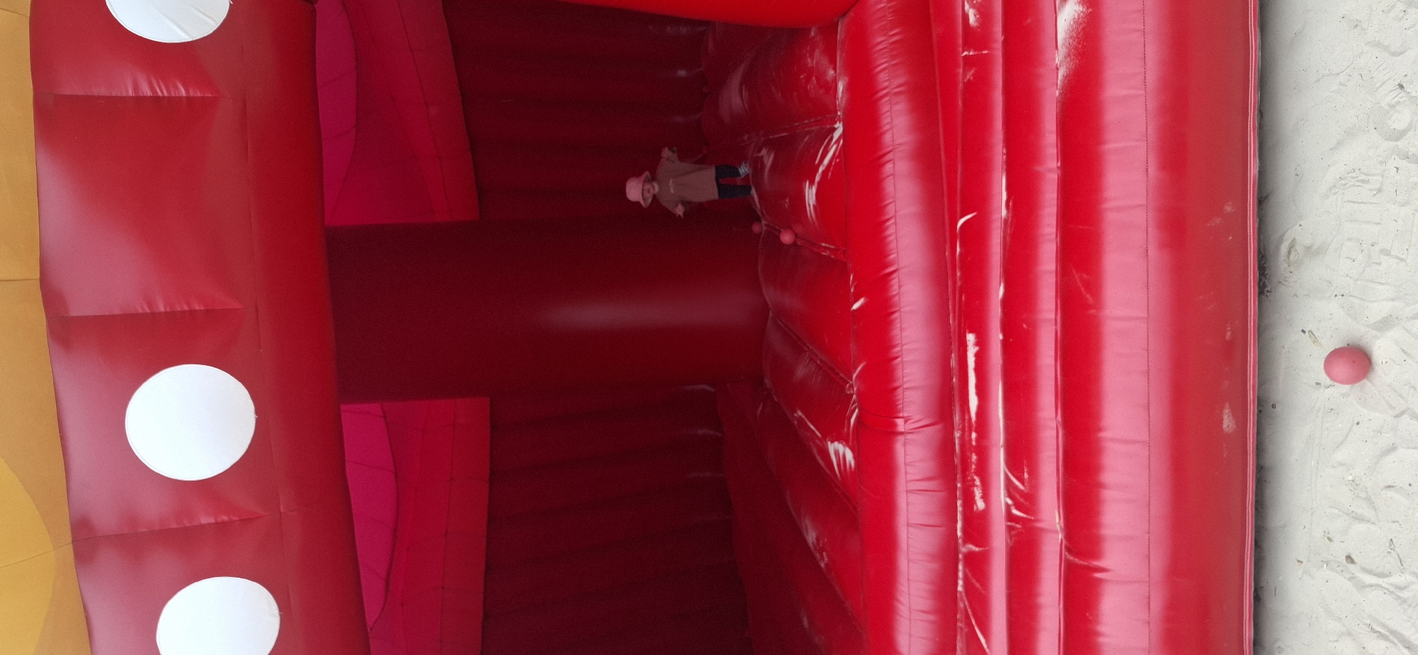 In Hürth, Germany, a young child wearing a beige bucket hat and a loose beige shirt with black pants stands on a red inflatable structure. The structure has a large central red pillar and is covered in inflated red sections. The top of the inflatable structure features three white circles on a red surface, with a hint of yellow above. There are several small pink balls scattered on the red inflatable surface and one on the sand below. The scene appears to be outdoors on a sandy surface.
