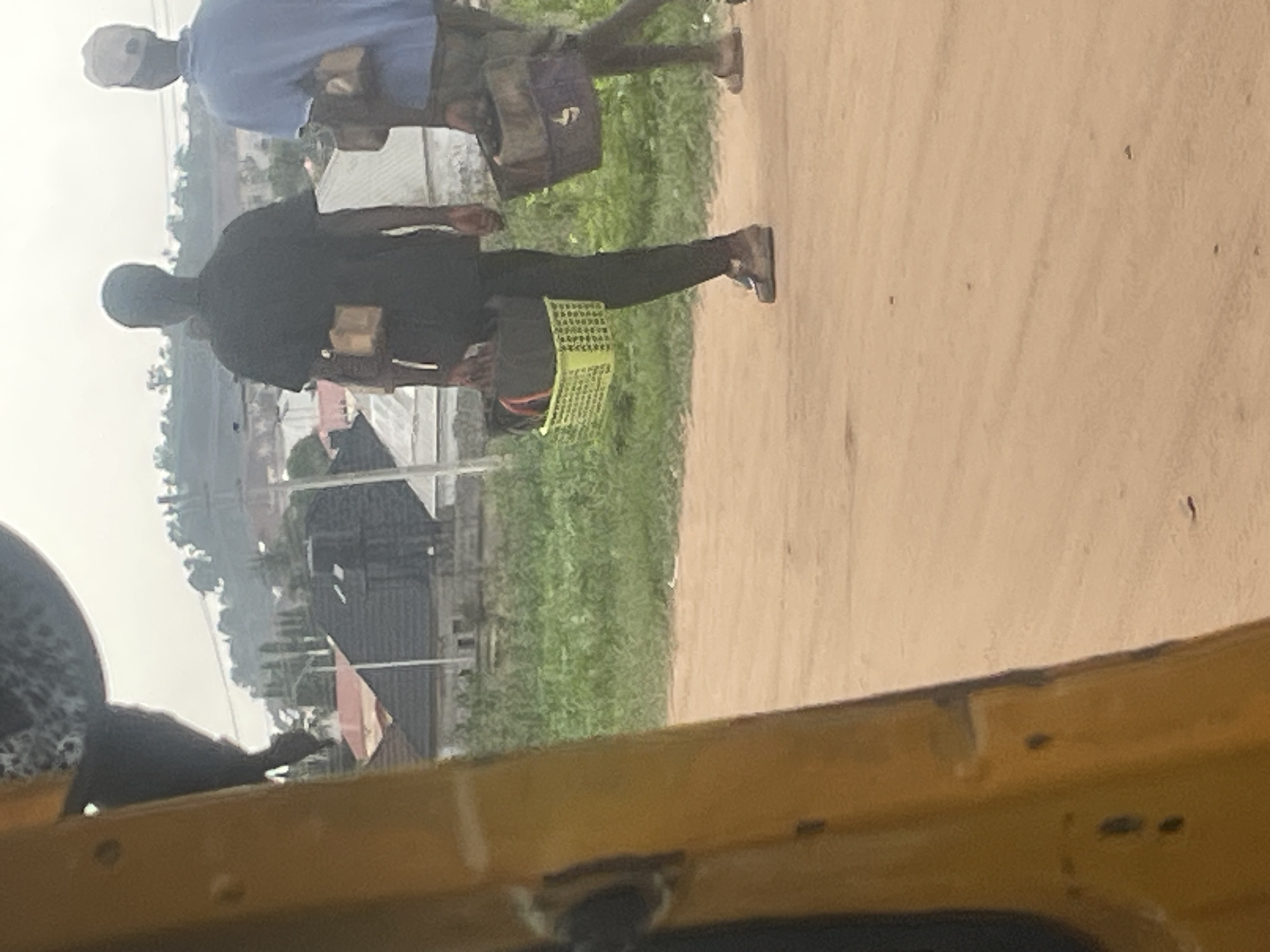 In Galadimawa Village, Nigeria, two individuals walk along a dusty road. The person in the foreground, wearing a black t-shirt and dark trousers, carries a lime green plastic basket filled with dark bags. They are also wearing brown shoes. To their right, a second person, dressed in a blue t-shirt and khaki trousers, carries a dark bag over their shoulder. They are wearing sandals. In the background, residential buildings with dark roofs are visible, interspersed with green vegetation and a hazy sky. The foreground is dominated by the yellow structure of what appears to be a vehicle.