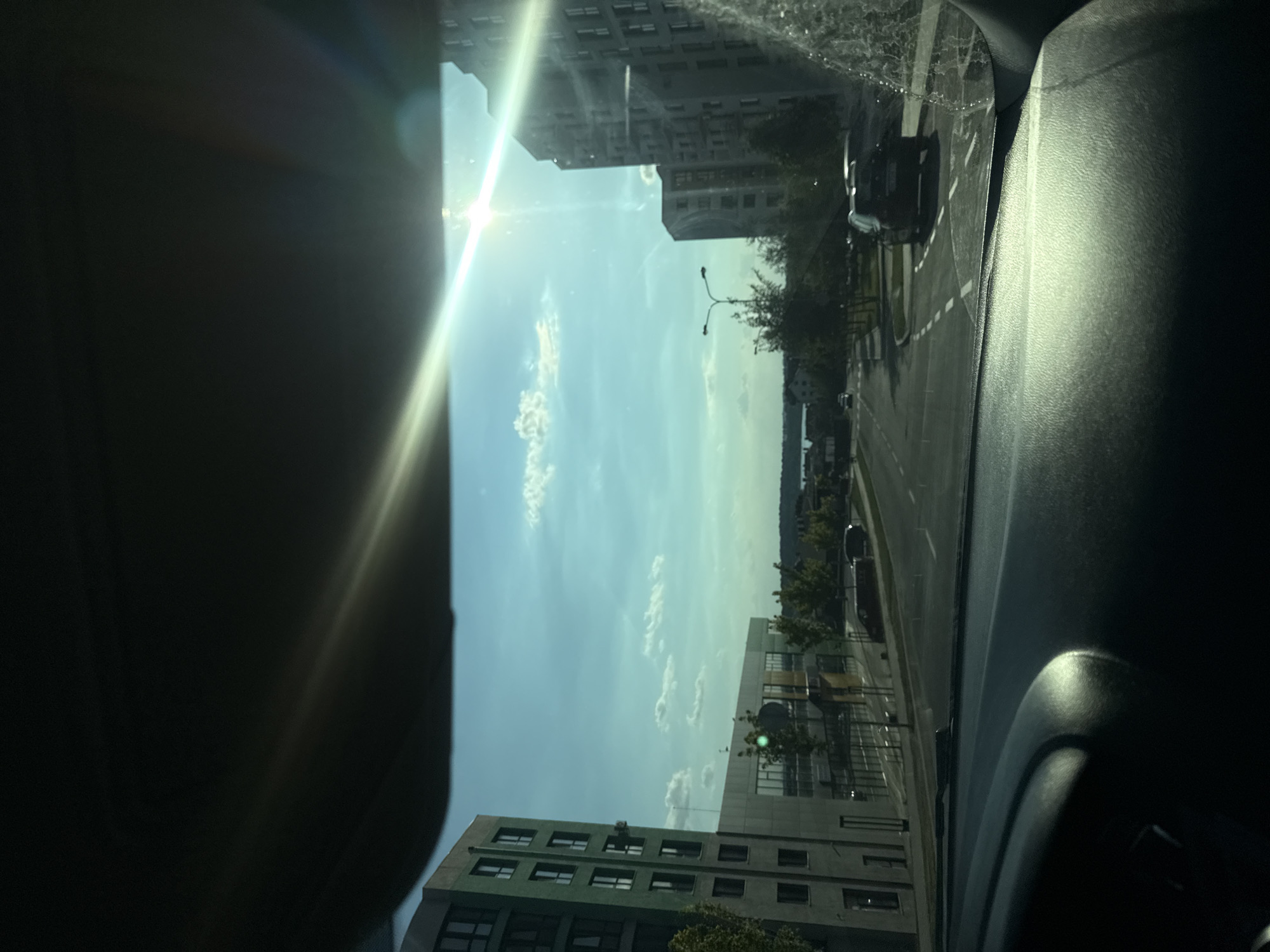 This is a point-of-view shot from inside a car looking out the windshield at a sun-drenched city street. The sun is bright and flares across the windshield, obscuring some details. The scene shows a road with a few parked cars, including a dark SUV on the right. Several modern buildings with multiple stories line the street, some appearing to be residential or office complexes. Trees and greenery are visible between the buildings and along the roadside. In the distance, there is a body of water, suggesting the city is near a lake or river. The sky is blue with scattered clouds. The interior of the car, specifically the dashboard, is visible in the foreground, with a sun glare reflecting off its surface.