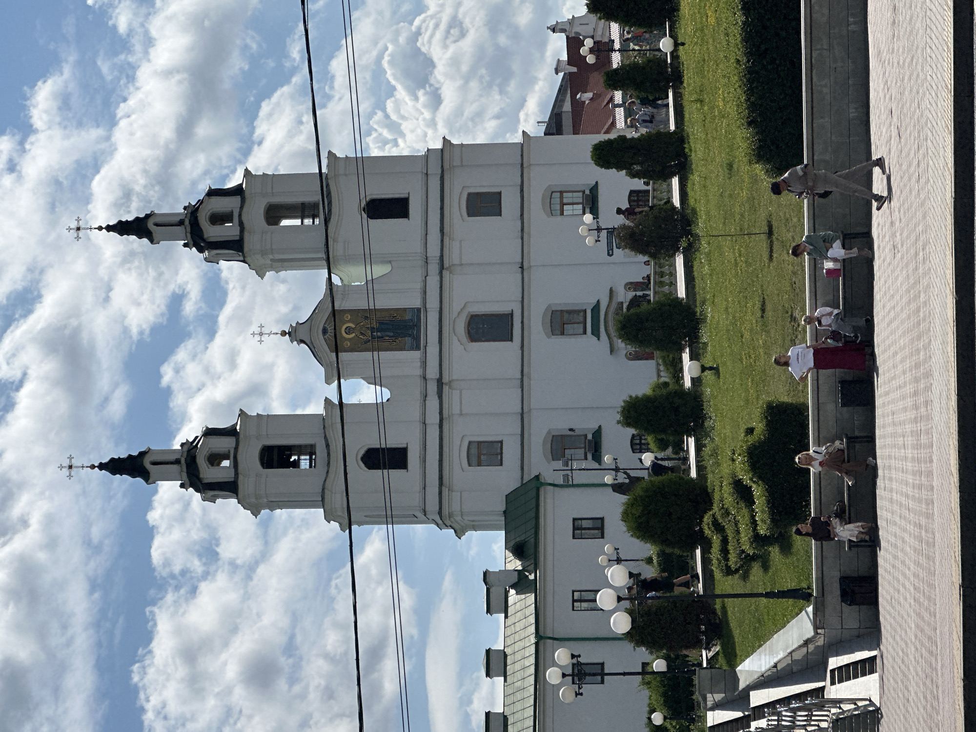 The image shows a frontal view of the Holy Spirit Cathedral, a prominent landmark in Minsk, Belarus. The cathedral is a large, white building with two prominent bell towers topped with black spires and crosses. Above the entrance between the towers is a mosaic of a religious figure. The sky is blue with scattered white clouds.
In the foreground, there is a paved walkway with several people sitting on benches and walking. One person is walking away from the camera towards the right side of the image. Another person, a woman in a red skirt and white top, stands near the edge of the walkway. Behind the walkway is a well-maintained grassy area with manicured bushes and trees. Several lampposts with multiple globe-shaped lights are visible along the walkway and on the grassy area. To the left of the cathedral, a section of a building with a green roof is visible. The overall scene depicts a public space in front of a significant religious and architectural monument.