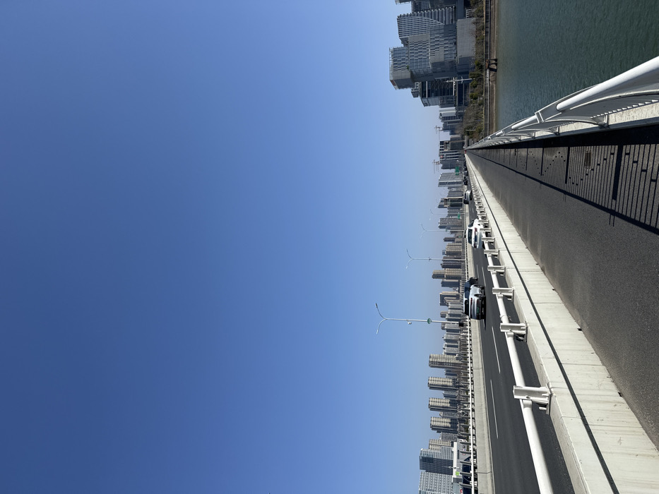 An expansive blue sky occupies the upper left half of the image. To the right, a long bridge extends into the distance, lined with skyscrapers on both sides. A line of cars moves along the bridge. The right edge features a curved white railing and dark shadow, suggesting a pedestrian walkway. The buildings near the far end of the bridge abut a body of water.