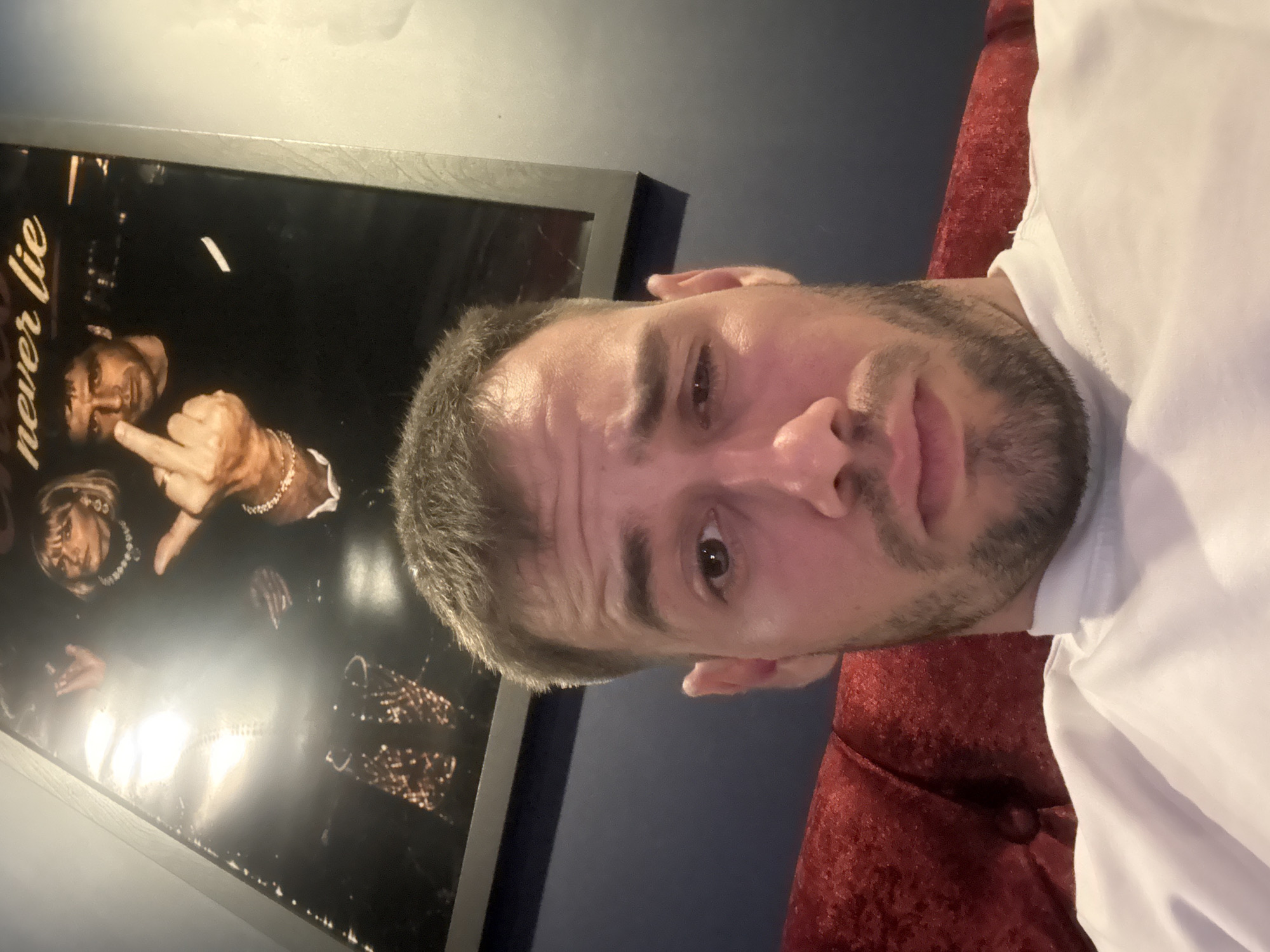 A man with short, dark hair and a trimmed beard is centered in the foreground, wearing a white shirt. Behind him on the left is a framed poster featuring an individual extending their middle finger. The background includes a dark wall and a portion of what appears to be a red, textured chair or sofa. There is nothing in the image that indicates a location in the city of Burgas, Bulgaria.