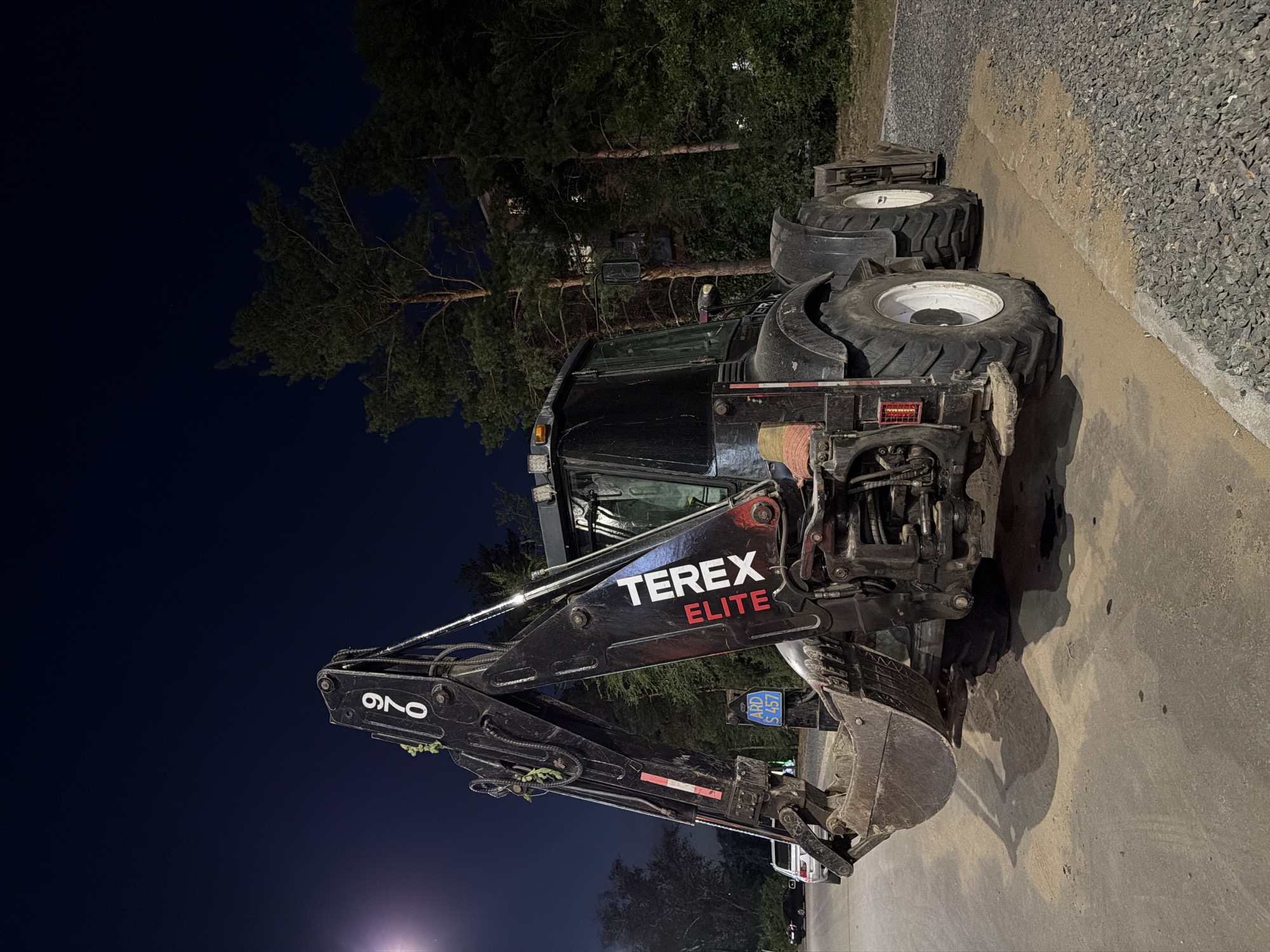 A black Terex Elite backhoe loader is parked on a gravel and asphalt surface at night. The excavator's arm is raised, and its bucket is lowered to the ground. The machine has the number "970" printed on its arm. A blue license plate with white lettering is visible on the side of the machine. The background consists of trees and a building. The scene is lit by artificial light, creating shadows. The location is specified as the city of Pavlodar, Kazakhstan. There are no people visible in the image.