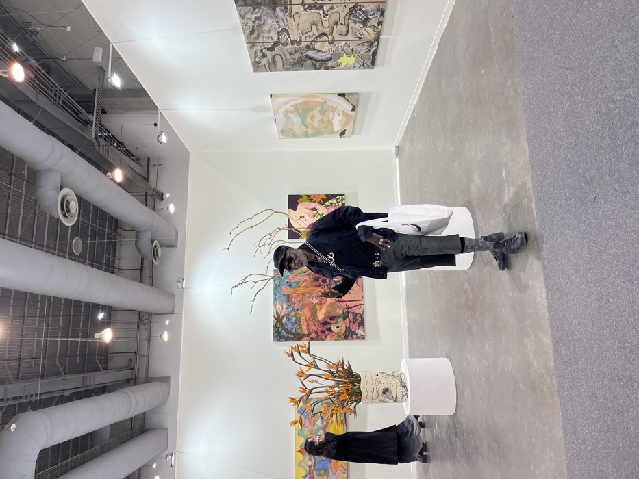 An art gallery displays vibrant paintings and a floral arrangement on white plinths. Overhead, exposed industrial piping and lighting fixtures create a modern atmosphere. A person in a black jacket, cap, and sunglasses stands near the artwork, holding a white tote bag. Another individual, wearing black, appears to be observing the floral piece. The flooring transitions from a polished concrete look to a dark carpet, defining the gallery space.