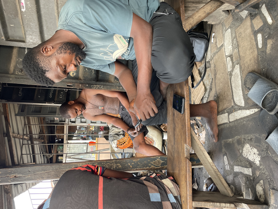 Two men are sitting on a wooden bench. One man wears a light blue shirt with a Lacoste logo and dark pants. The other man is shirtless and wears dark shorts. The man in the shorts is cutting his toenails. A black smartphone and a black bag are on the bench next to the men. A tile floor is visible.