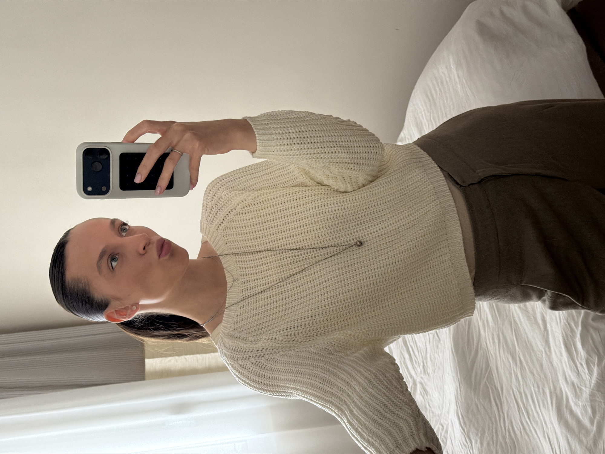 A woman is taking a selfie. She is wearing a cream-colored, knit sweater, olive green pants, and a silver necklace. She is holding a phone with a light gray case. She has dark hair pulled back and is making a slightly pursed lip expression. The background appears to be a bed with white sheets. There is no information to indicate the location is in Budva, Montenegro.