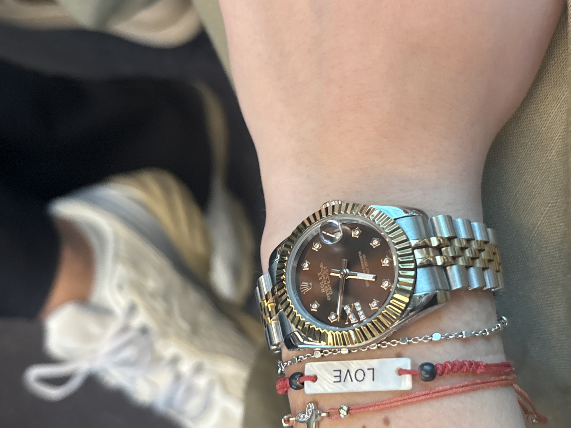 A person's wrist is shown wearing a two-tone stainless steel and gold Rolex Datejust watch with a brown dial and diamond hour markers. The watch is worn above several bracelets: a silver chain, a red braided string bracelet with a white tag that says "LOVE," and a red string bracelet with a silver cross charm. Part of a white and gold sneaker is visible in the background. The person is wearing a green jacket. There is no information in the image that references the city of Alessandria, Italy.