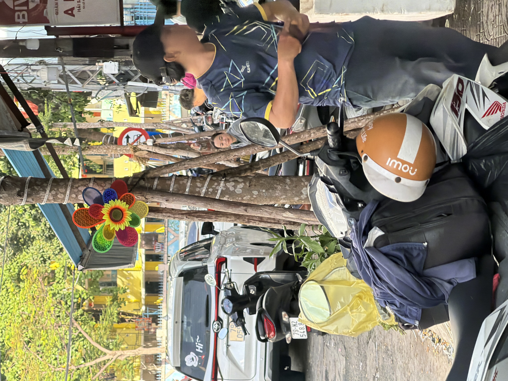 In Nha Trang Ward, Vietnam, a street scene unfolds with several elements. In the foreground, a motorcycle is parked, partially obscured by a yellow plastic cover and a dark blue jacket draped over its seat. A white and orange helmet rests on the seat. To the left, a white SUV with a "Hi~" sticker featuring Mickey Mouse is parked on a paved surface. Behind it, another black scooter is visible. The background reveals a yellow building with trees and foliage. Several trees with textured bark are positioned throughout the scene, with one serving as a vertical support for a colorful pinwheel. A red and white circular sign with an arrow pointing left is attached to one of the trees. Several individuals are present: a person wearing a black baseball cap and a dark blue t-shirt with yellow and blue geometric patterns stands in the mid-ground, looking to the right. Another person, a woman with dark hair, is seated further back, looking towards the left. The environment appears to be a street or sidewalk with parked vehicles and overhead structures.