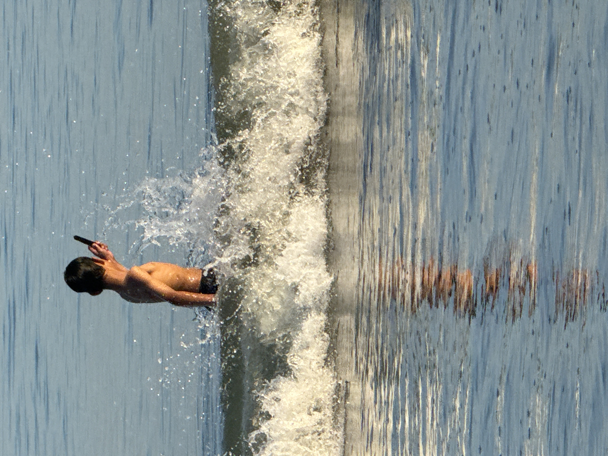 The image, taken in Jimbaran, Indonesia, depicts a shirtless individual, likely a young male, holding a black object to his head. He is positioned horizontally in the frame, seemingly suspended mid-air as if he is diving. Splashes of water surround him. The background consists of what appears to be a vertically-oriented water surface, possibly the ocean, with distorted reflections visible.