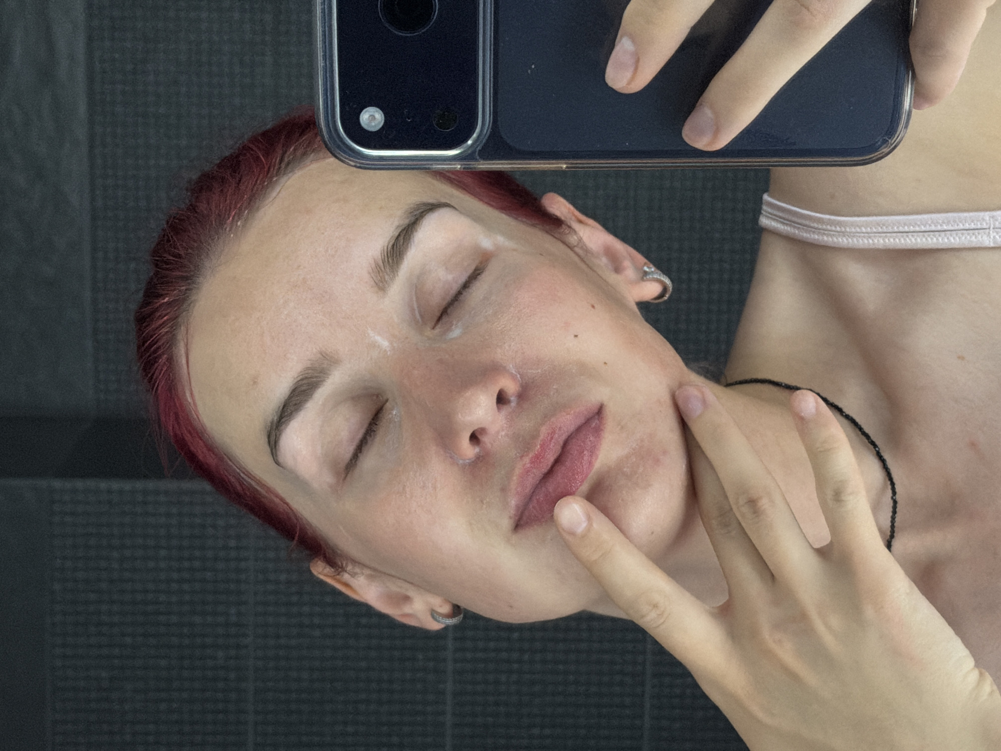 A young person with red hair is taking a selfie in a bathroom. They are looking down with their eyes closed and one hand touching their lips. The person is wearing a light-colored camisole. The background is a dark, textured wall. There are no specific identifiable landmarks or objects that would indicate the location within the city of Chaikovsky, Russia.