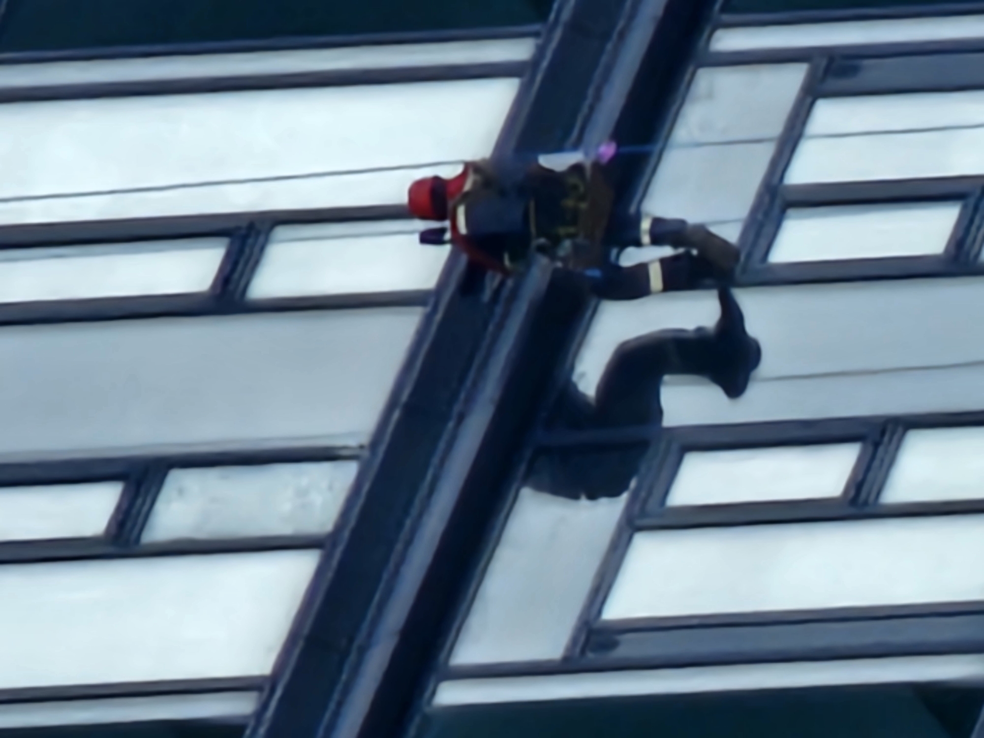 A person is rappelling down the side of a modern building, characterized by its large, reflective glass windows and dark framing. The individual is wearing a red helmet and dark clothing with what appears to be some yellow accents on the legs. They are suspended by ropes and are equipped with a harness and safety gear. The building is tall, with multiple stories visible, and the windows reflect the sky and surrounding environment, giving it a slightly abstract appearance. The scene suggests a high-rise window cleaning or maintenance operation taking place on a clear day. The location is identified as Moscow, Russia.
