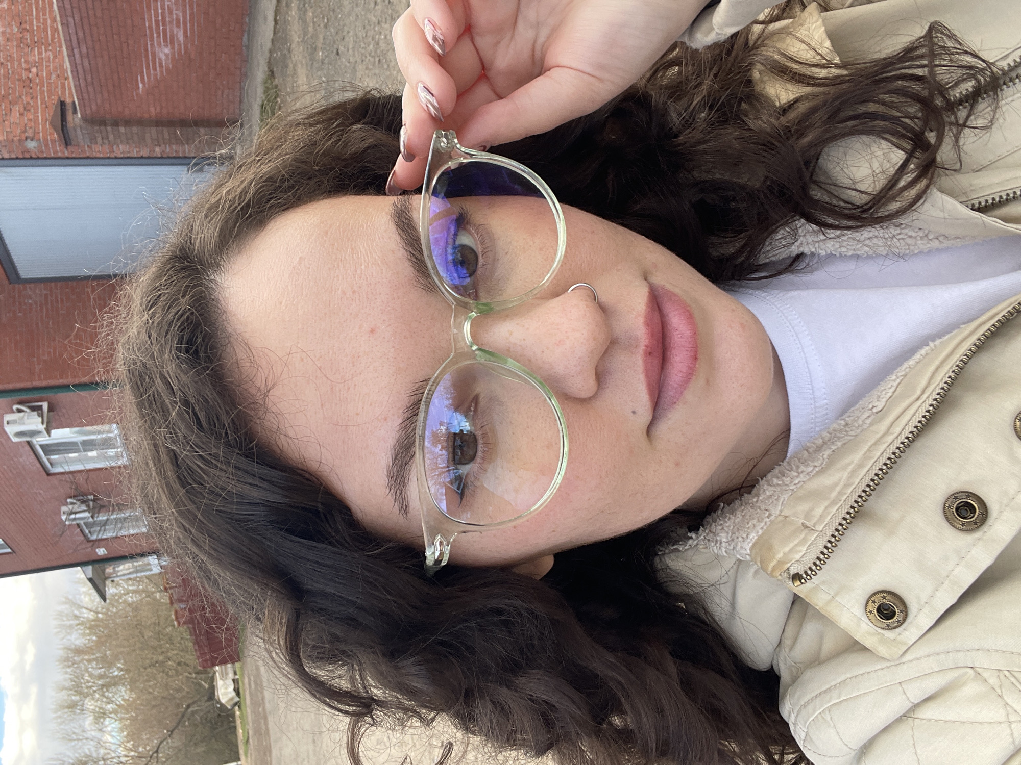 A close-up outdoor portrait features a young woman with dark, curly hair. The image is taken at a slight upward angle, with the woman looking directly at the camera. She is wearing light-framed glasses, which she is holding with her right hand by the upper rim. Her fingernails are painted with a shimmery polish. A silver ring pierces her nose. Her lips are a light pink or nude color, slightly smudged.
She is dressed in a light-colored, possibly beige or cream, jacket with a textured, sherpa-like collar and a visible zipper and metal snap buttons. A white t-shirt is visible underneath.
The background suggests an urban outdoor setting under an overcast sky. To her left, a reddish-brown brick building with windows and air conditioning units is visible. Further in the background, bare trees indicate a cooler season. The ground appears to be a paved or dirt path. There is no legible text visible in the image. The overall impression is a casual, everyday moment captured in what appears to be Izhevsk, Russia.