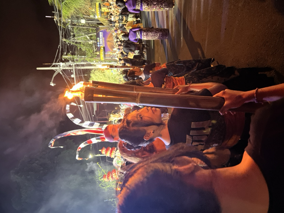 This image captures a nighttime ceremonial procession on a street in Munggu, Indonesia. In the foreground, the blurred left shoulder and back of an individual holding a bamboo torch are visible. Immediately to their right, a young woman with dark hair styled in two buns, wearing a black t-shirt with a light logo and a red sash over a dark patterned sarong, faces right, holding three flaming bamboo torches that emit smoke. Behind her, another participant in an orange top and patterned sarong also carries a torch. Further back, a line of individuals dressed in purple tops and dark patterned sarongs walks away from the viewer. To the left, several tall, curved ceremonial poles with red, white, and black striped fabric, and ornate tiered structures are visible. In the background, a large crowd gathers under artificial lighting, with some people in traditional attire. A prominent banner displays "OOOO GOGO OPEN" over an illuminated graphic of a bridge. Overhead utility lines and streetlights are visible against the dark sky.