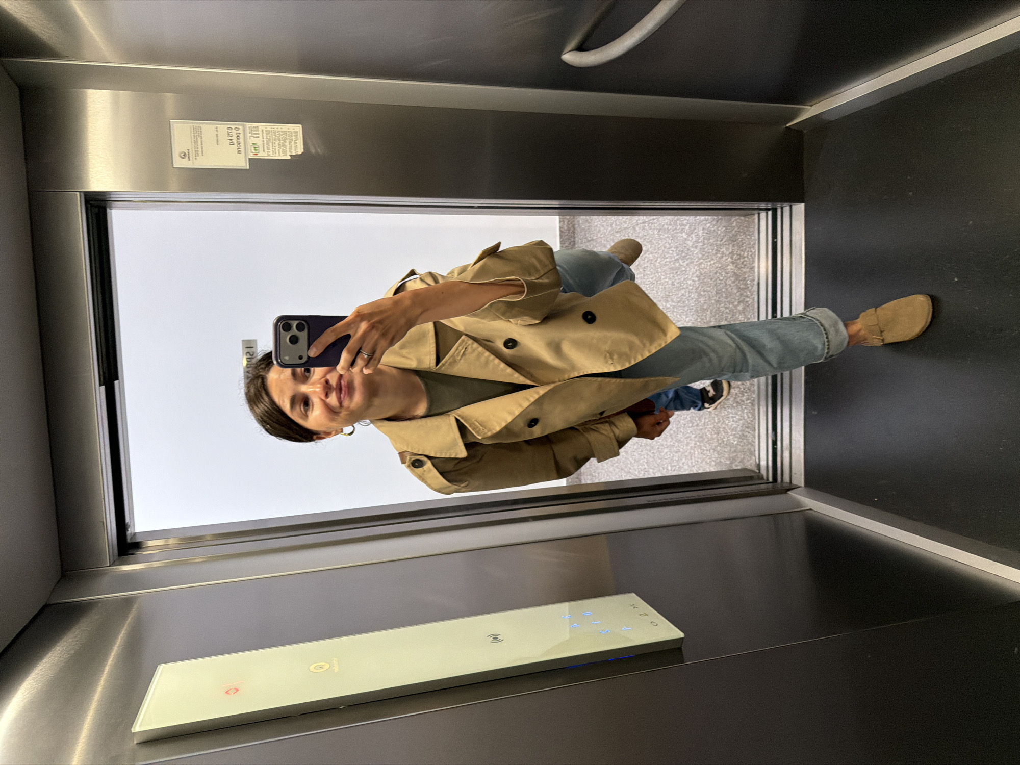 A woman takes a selfie in an elevator. She wears a khaki trench coat, light blue jeans, and tan shoes. The elevator interior is gray metal with a white button panel featuring blue lights. Through the elevator doors is a glimpse of Budva, Montenegro.