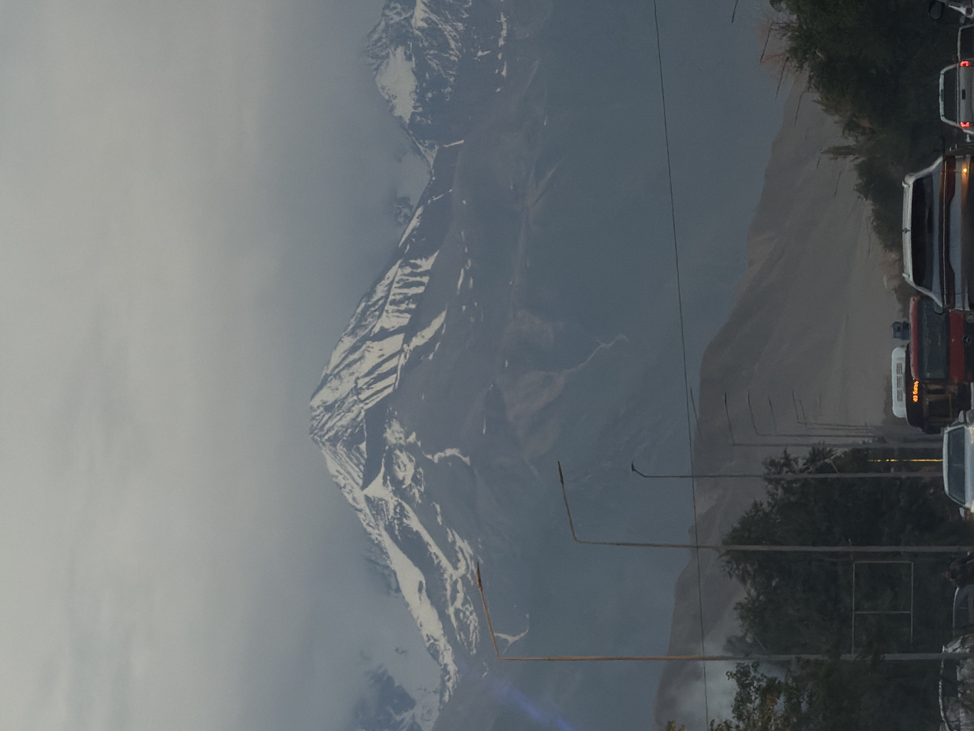 The image depicts a scenic view of a snow-capped mountain range under a cloudy sky. In the foreground, a road with multiple lanes is visible, with cars driving on it. Streetlights line the road, and trees and hills are present on either side. The overall atmosphere appears to be overcast, possibly indicating dawn, dusk, or a cloudy day. The image is taken from within a vehicle, suggesting a perspective from a moving car or a traffic jam.