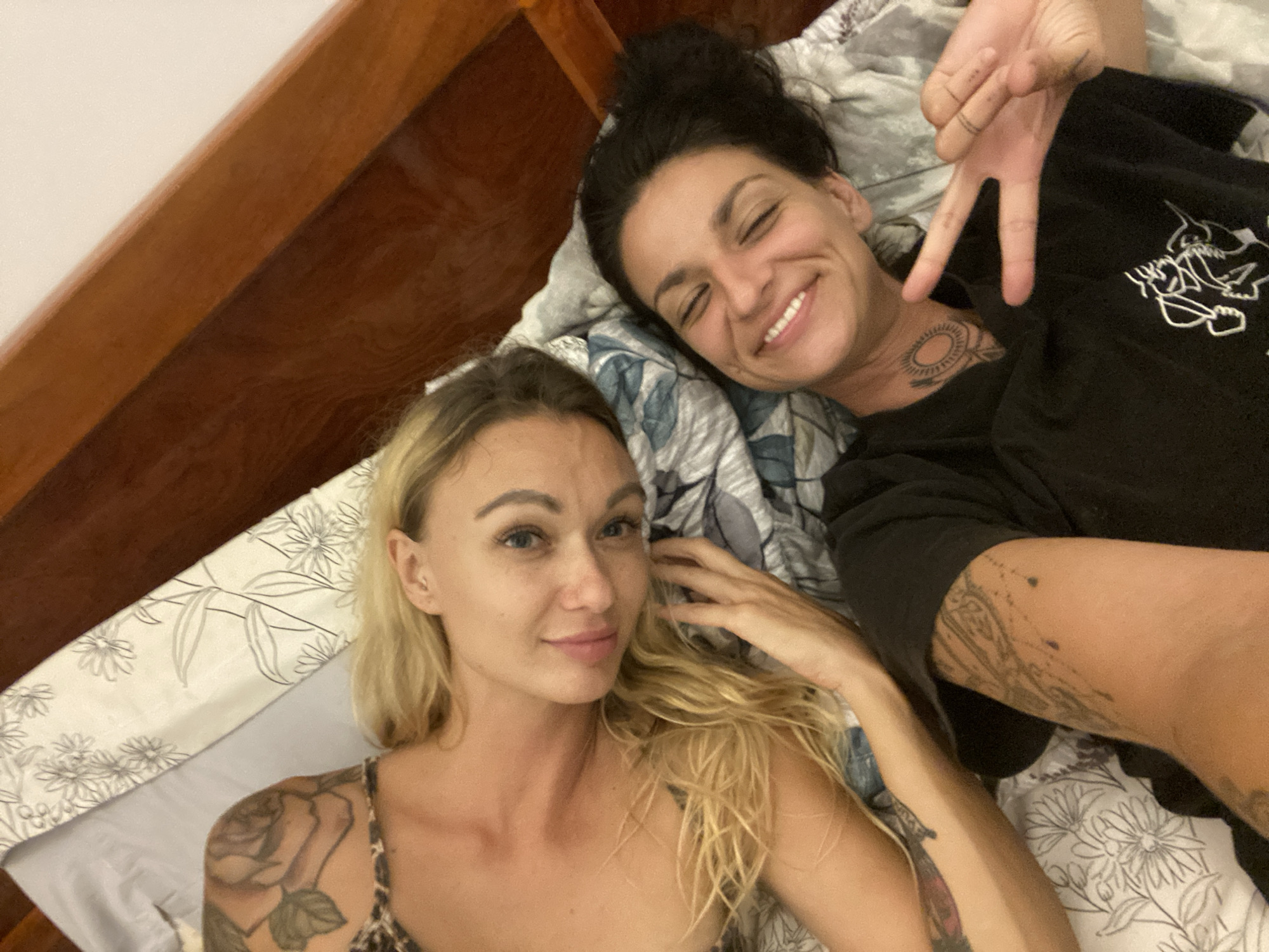 Two individuals are lying on a bed, indoors, in Mũi Né, Vietnam. The person on the left has blonde hair and is wearing a leopard print top, with visible tattoos on their arm and shoulder. The person on the right has dark hair and is smiling, wearing a black t-shirt with a graphic print. They are making a peace sign with their fingers. The bed has floral patterned sheets and is positioned next to a wooden headboard.