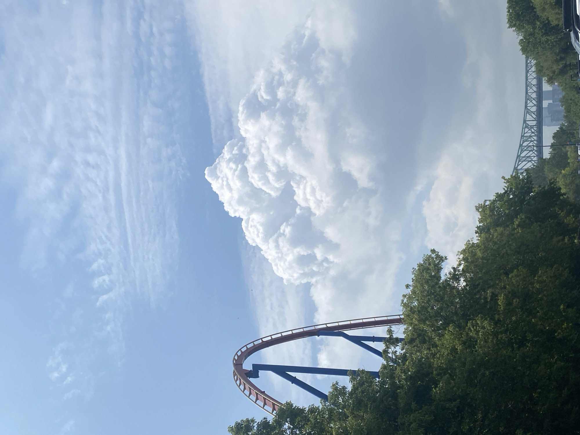 This is a vertical, eye-level, outdoor photograph of a roller coaster track and a large cloud formation on a bright, partially cloudy day in Montreal, Canada. The roller coaster, painted red and blue, is in the left half of the image, curving upwards against the sky. Lush green trees fill the lower left portion of the frame, partially obscuring the roller coaster's support structure. Dominating the center and right of the image is a massive, white, cumulus cloud, its billowy texture clearly visible. In the far distance, a blue metal truss bridge spans a body of water, with buildings of the city skyline visible beyond it. The sky is a bright blue, with wispy cirrus clouds scattered above the larger cumulus formation. There are no people or animals visible in the frame, and no discernible actions or interactions are taking place. The overall impression is one of a sunny day at an amusement park with a prominent cityscape in the background.