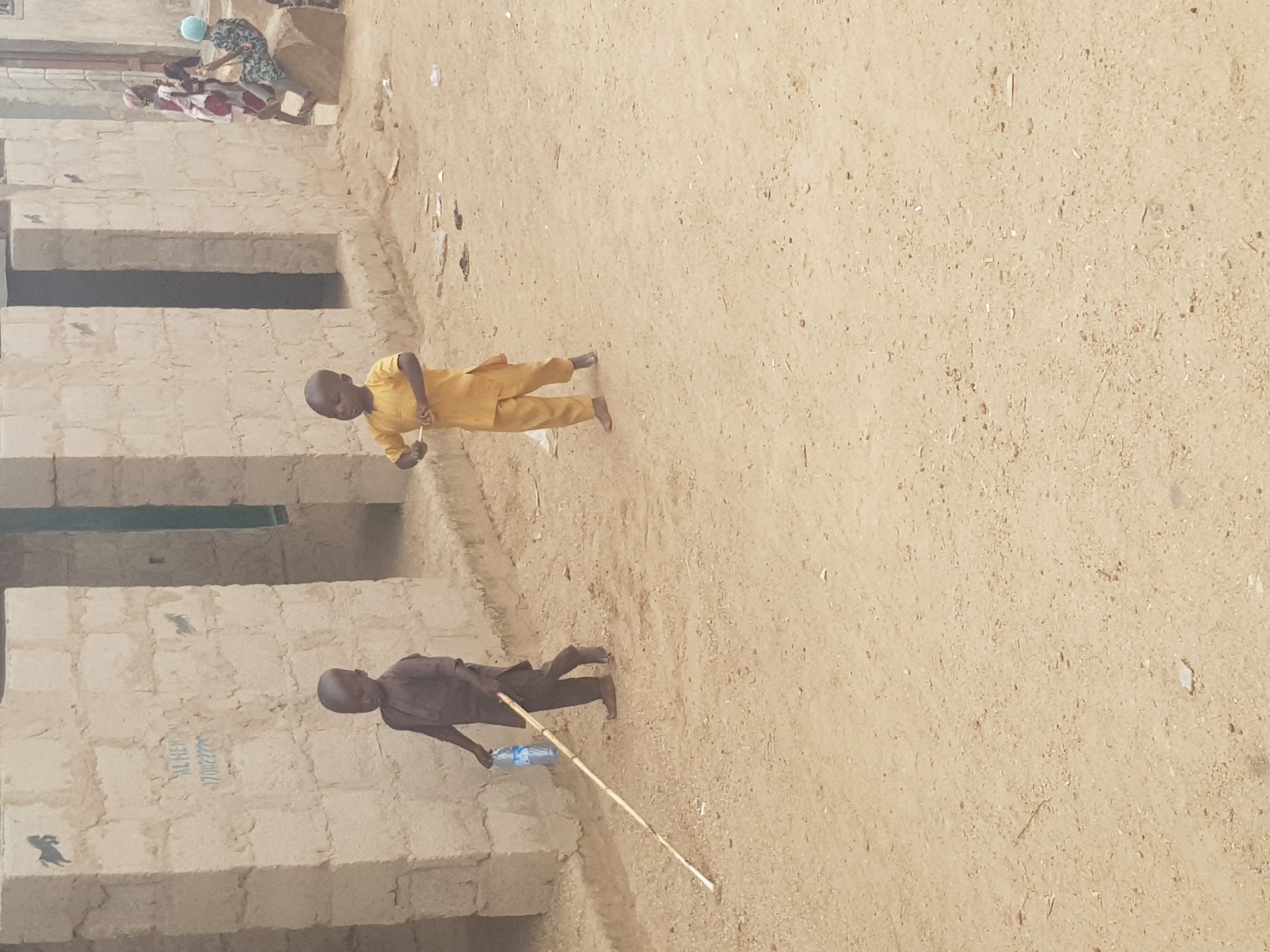 The image depicts an outdoor scene in Dutsinma B, Nigeria. Two young boys are present. One, clad in a yellow outfit, stands on the dirt ground. The other, wearing a darker outfit, stands near the edge of a rough concrete structure holding a stick and a water bottle. In the background, a group of people are clustered near a building. The ground is sandy and sparsely littered.