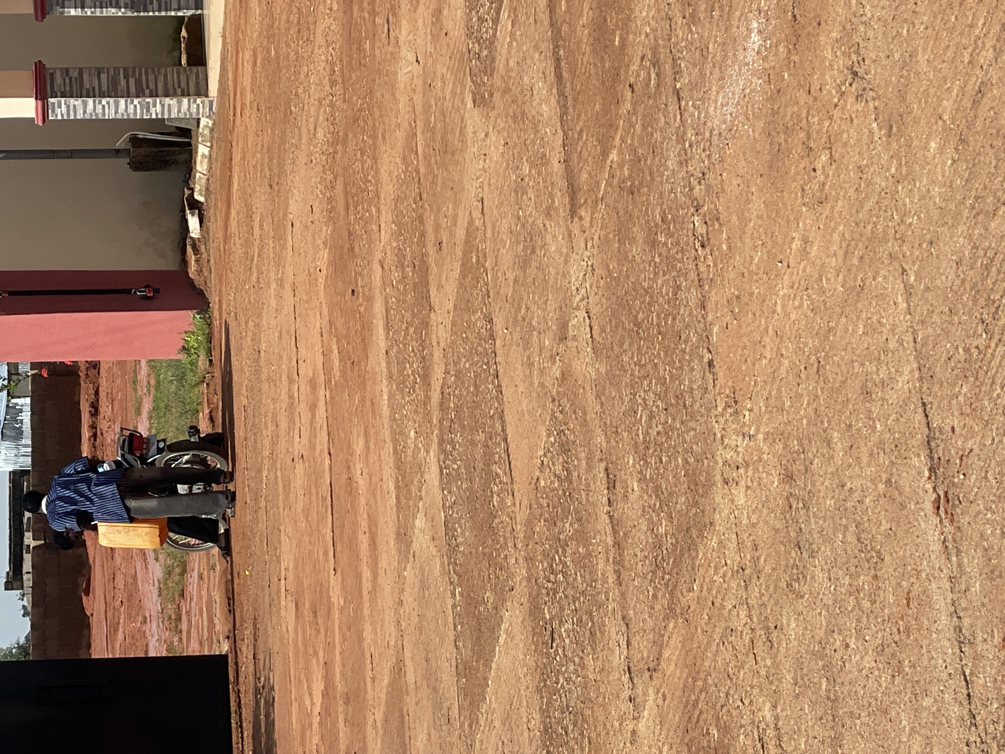 In a dusty, outdoor area in Jos, Nigeria, a man wearing a blue and white striped shirt and dark pants stands next to a motorcycle. He is holding a large, yellow jerrycan, which appears to be full. The motorcycle's rear wheel is visible. The ground is covered in reddish-brown dirt, with some indications of paving stones. In the background, there are buildings under construction, including a wall made of brick and corrugated metal roofs. A bright pink pillar is also visible, suggesting this is an entrance or part of a structure. The scene appears to be taking place during the day, with a clear sky.