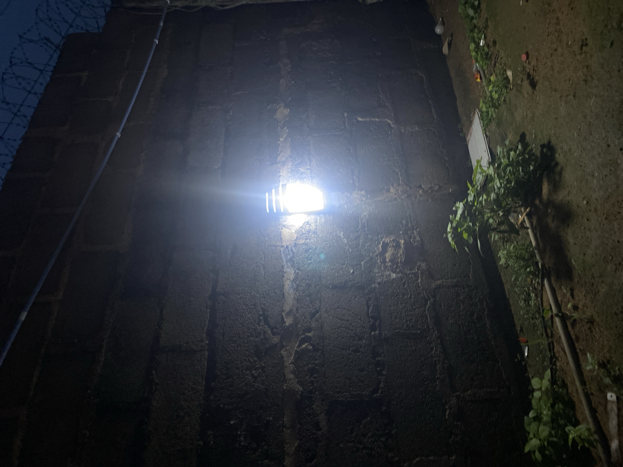 The image captures a nighttime scene featuring a textured, dark brick or block wall illuminated by a single, intensely bright white light source. The light, likely an outdoor security or utility lamp, is mounted on the wall and causes significant lens flare, creating a strong beam of light that extends outwards.
To the left of the wall, a security feature resembling barbed or razor wire is visible against a dark, possibly twilight sky. A light-colored pipe or electrical conduit runs vertically along the left side of the wall. To the right, the ground is visible, appearing dark and earthy, with scattered green vegetation, including some small bushes or plants growing near the base of the wall. Some light-colored debris is also discernible on the ground.
The overall impression is one of a functional, possibly industrial or perimeter area, at night. There are no people visible, and no specific activity is taking place. The scene is static and provides a glimpse of a secured boundary under artificial illumination. No text is visible in the image. The time of day is clearly night due to the darkness and the reliance on artificial light. The location context provided is Du, Nigeria.