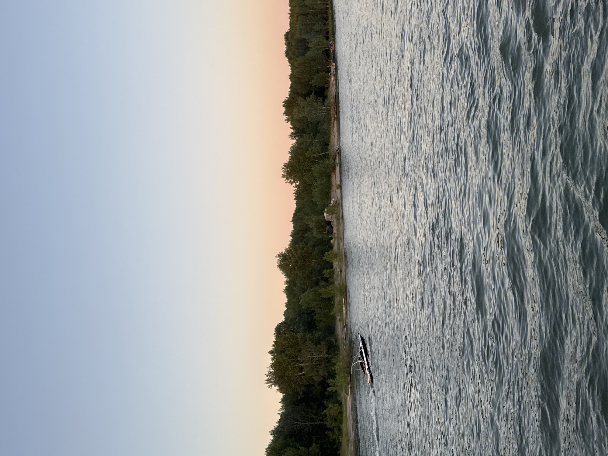 This media file depicts a serene lake scene at dusk, with a warm, gradient sky transitioning from a soft peach to a pale blue. The horizon is lined with a dense, dark green forest. The lake itself is calm, with gentle ripples catching the fading light. A single, black and white stand-up paddleboard is visible on the water, its wake suggesting recent movement. The overall atmosphere is tranquil and peaceful, evoking a sense of calm end-of-day relaxation. The location is hinted to be Novosibirsk, Russia.