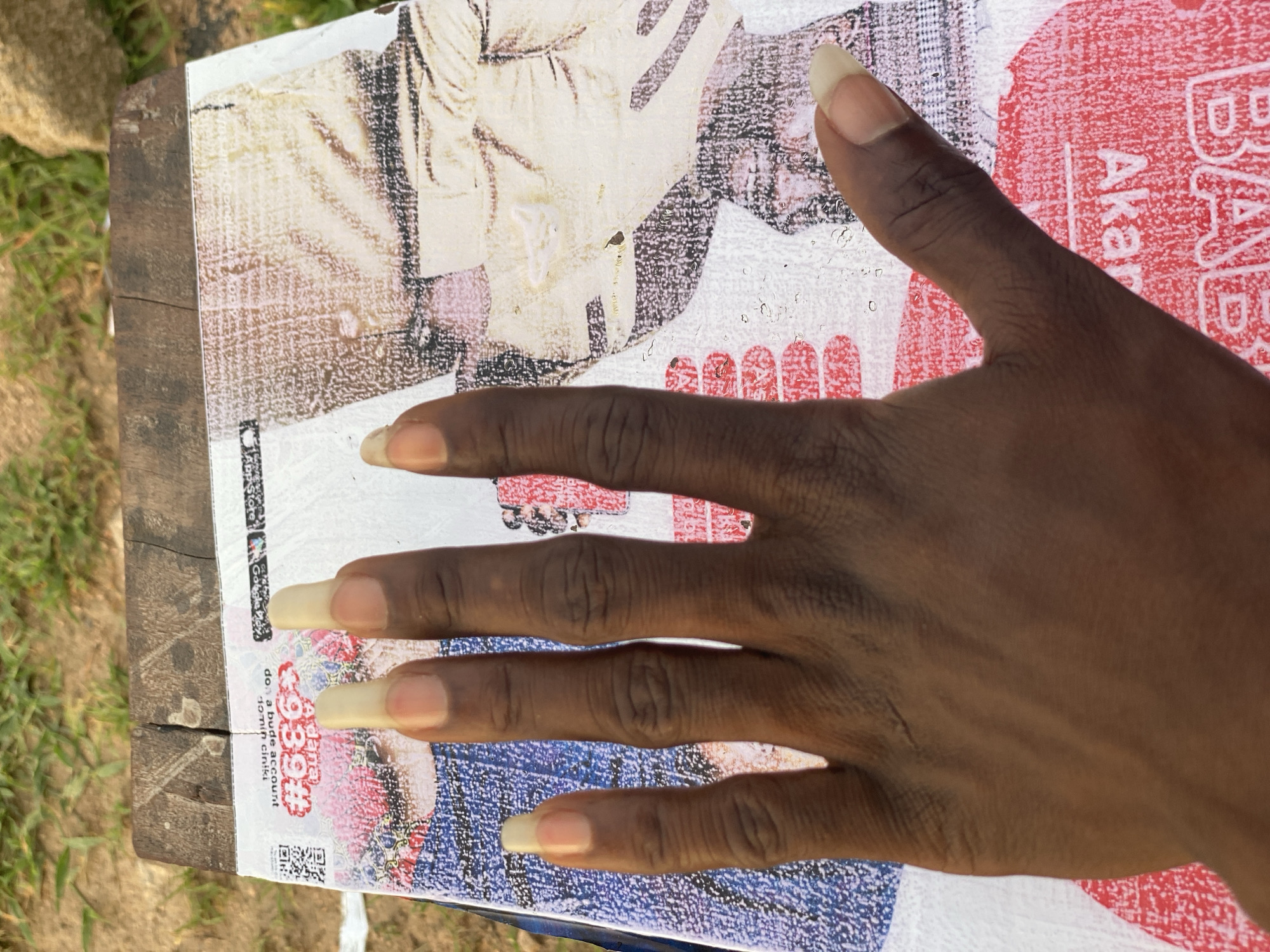 The image shows a close-up, overhead view of a left hand with long, light-colored fingernails resting on a piece of paper with printed text and images. The paper appears to be some kind of flyer or advertisement, with vibrant colors and graphics. The background consists of a rough, wooden surface, possibly a table or bench, and out-of-focus greenery and rocks, suggesting an outdoor setting. The lighting is natural and appears to be from the side, casting subtle shadows. There is no discernible location in Jalingo, Nigeria indicated within the image.