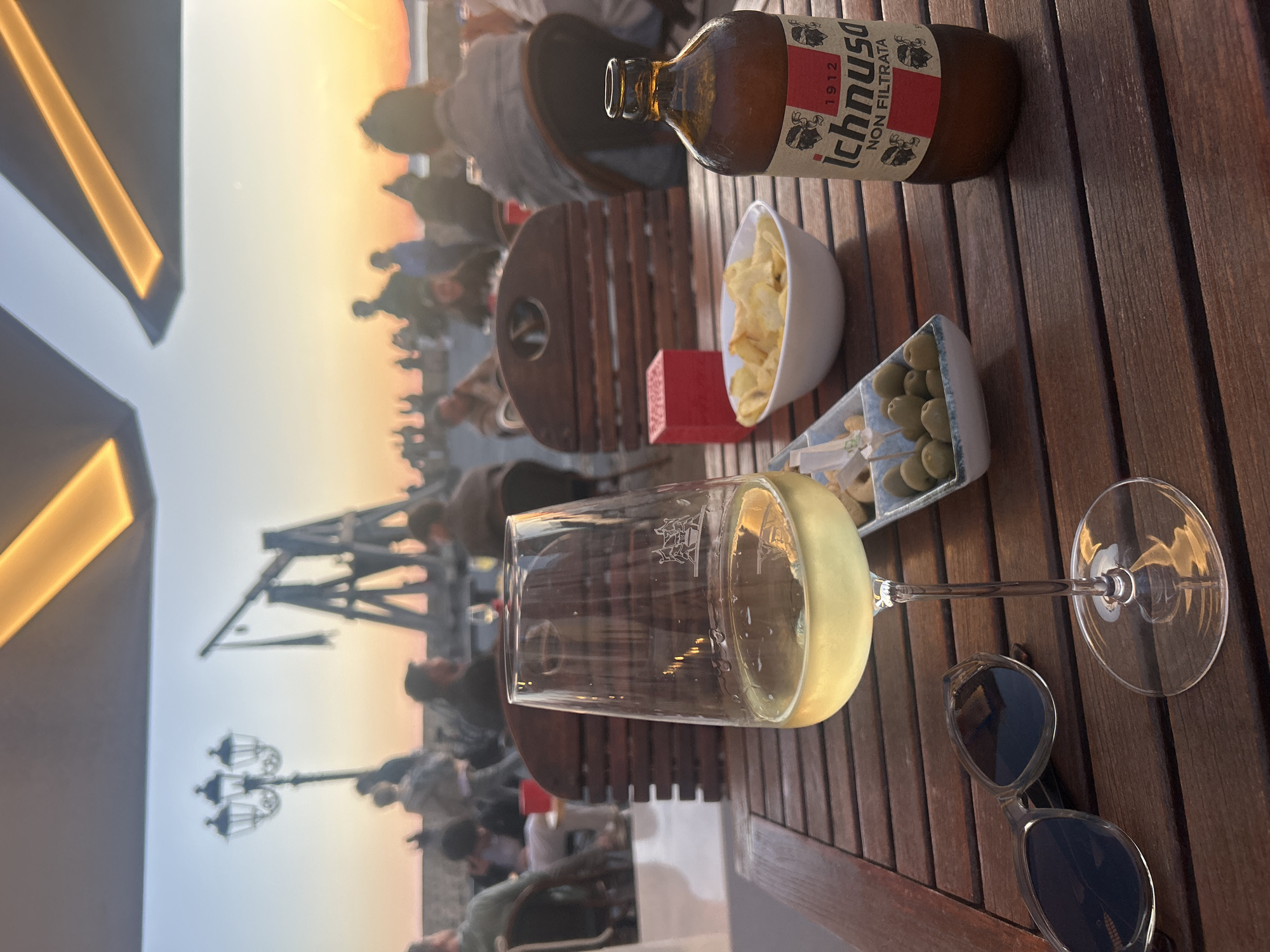 The image depicts a scene at sunset on a wooden table in what appears to be Alghero, Italy. The table holds a glass of white wine, a bottle of Ichnusa beer, a bowl of potato chips, a dish with olives and cheese cubes, and a pair of sunglasses. In the background, people are seen on a promenade or terrace, silhouetted against the sky which is illuminated by the setting sun. Also visible is a lamppost, and a crane-like structure, probably part of the harbor. The lighting suggests late afternoon or early evening, and the objects on the table suggest a leisurely dining or drinking experience. The image is taken at a slightly tilted angle.