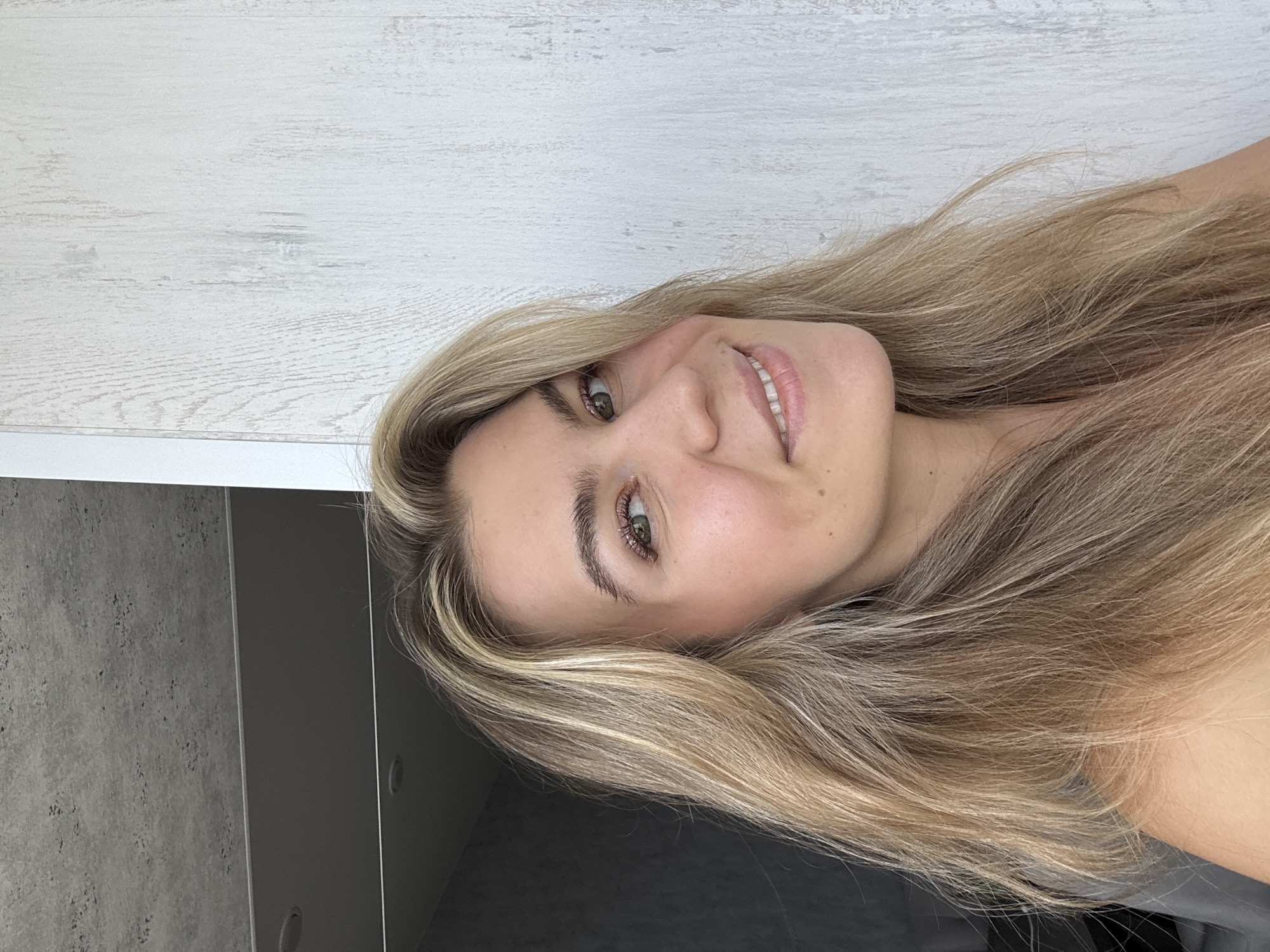 The image shows a woman with blonde hair and fair skin looking at the camera and smiling. The background consists of a white wall with a wood-like texture and a section of a dark grey cabinet. The location is unknown.