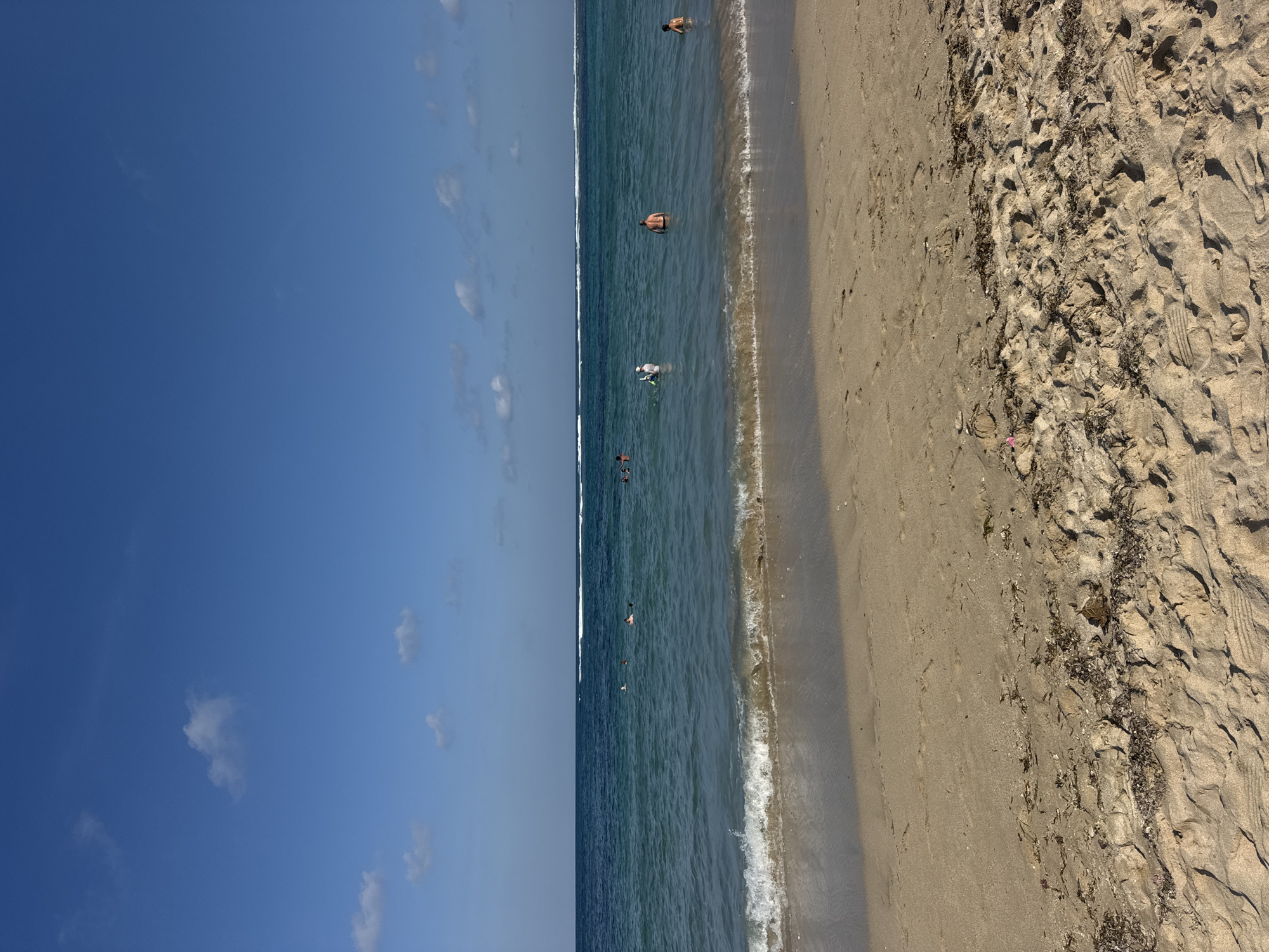 The image shows a sandy beach in Kuta Selatan, Indonesia, with the turquoise ocean in the background under a clear blue sky with scattered white clouds. Gentle waves are breaking on the shore. Several people are visible in the water, some swimming and others standing. The sand on the beach has scattered seaweed and footprints. The overall scene depicts a tranquil beach day.