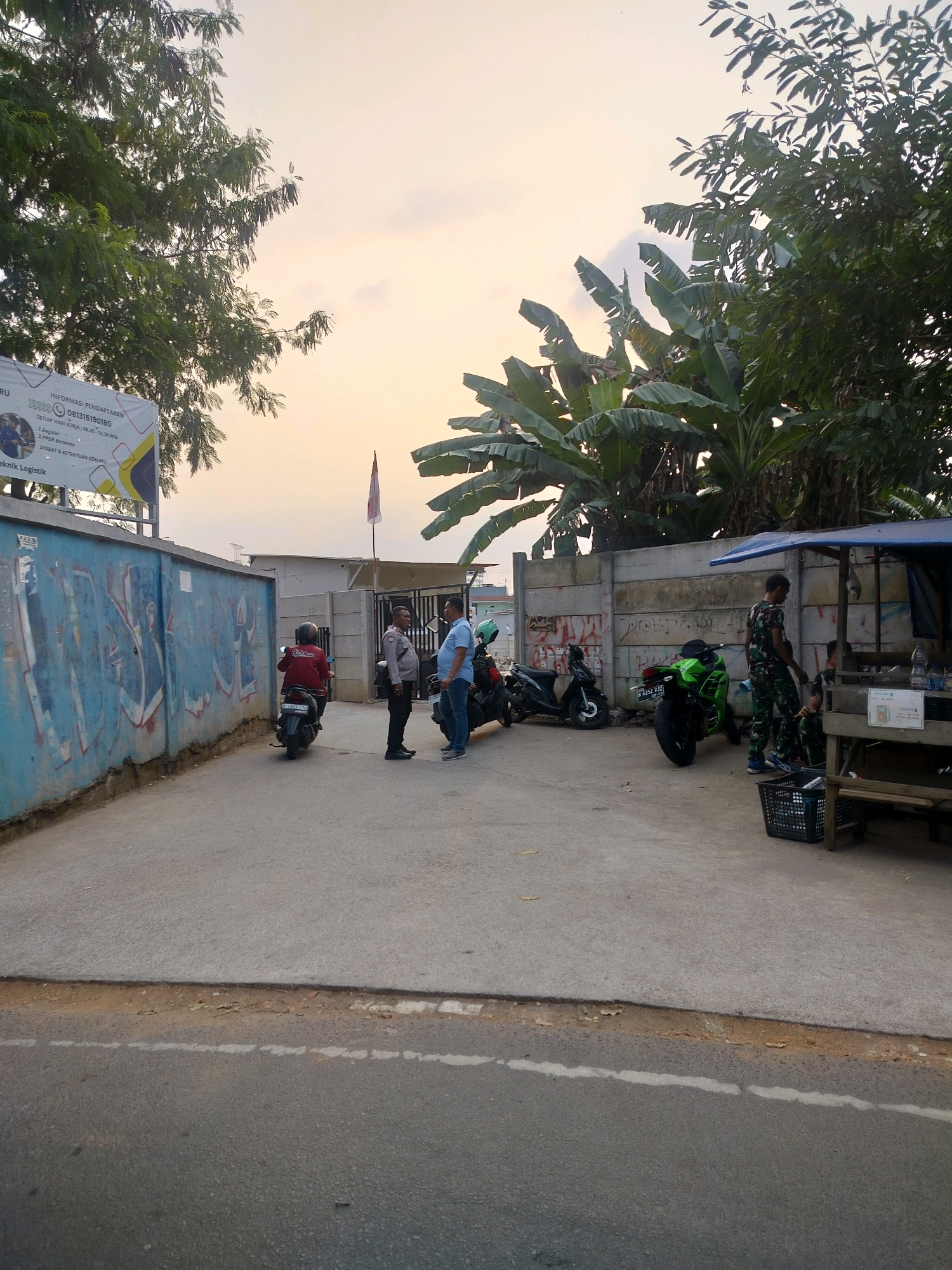 The image depicts an outdoor scene in an urban setting, possibly East Jakarta, Indonesia, given the context of the overall scene and tropical vegetation. The time of day appears to be late afternoon or early evening, with soft, warm lighting from the sky.
In the foreground, a paved area, likely a street or parking lot, transitions to a rougher, dirtier surface with a broken white line. Beyond this, a concrete walkway runs along a graffiti-covered wall.
A group of people are present. Two men in casual clothing are standing and conversing near a white gate. One man is wearing a blue shirt, and the other is in a dark uniform, possibly a police officer, with a badge visible on his chest. To their left, a person in a red jacket is riding a scooter, facing away from the camera.
Further to the right, there are several motorcycles parked. One is a bright green sportbike. A small roadside stall or kiosk with a blue tarp roof is set up, with a person in camouflage attire standing near it. Another person, possibly a child, is seated.
The background features concrete walls, some with graffiti, and a backdrop of lush green trees, including large banana trees, suggesting a tropical or subtropical climate. A flag is visible on a pole behind the gate. A banner with text and graphics is mounted on a pole to the left, providing information, likely about registration or services, with a phone number clearly displayed. The overall atmosphere is casual and everyday.