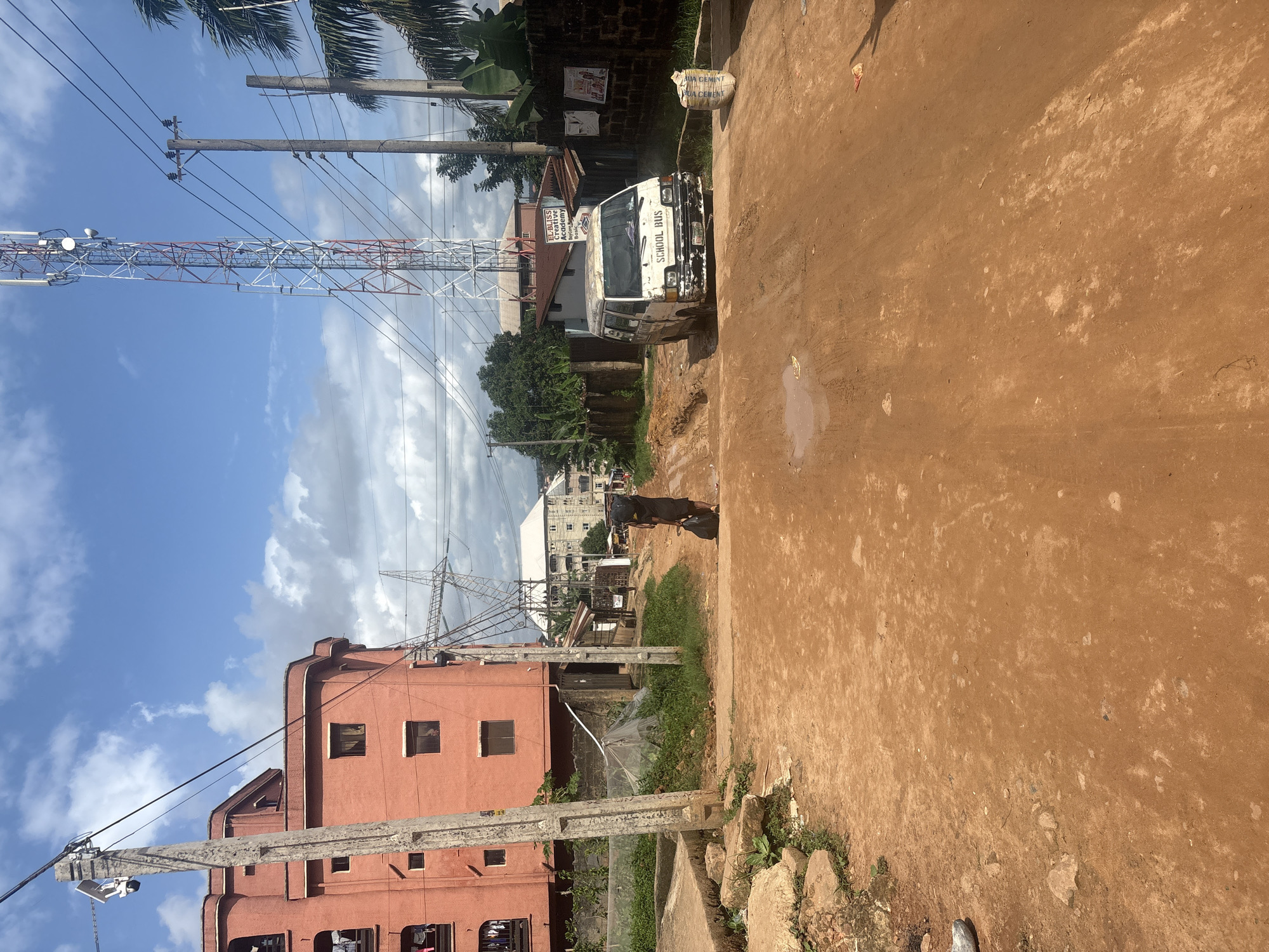 In Nkpor, Nigeria, a dirt road with muddy patches cuts through a residential area. A person carrying a large black bag walks away from the camera towards a white van parked in the middle of the road. The van, which appears to be a school bus, has "SCHOOL BUS" written on its front. To the left, a tall, orange building with balconies stands beside a concrete utility pole with electrical wires. In the background, a red and white telecommunications tower rises high into the sky, with other buildings and lush green trees visible. The sky is blue with scattered clouds.
