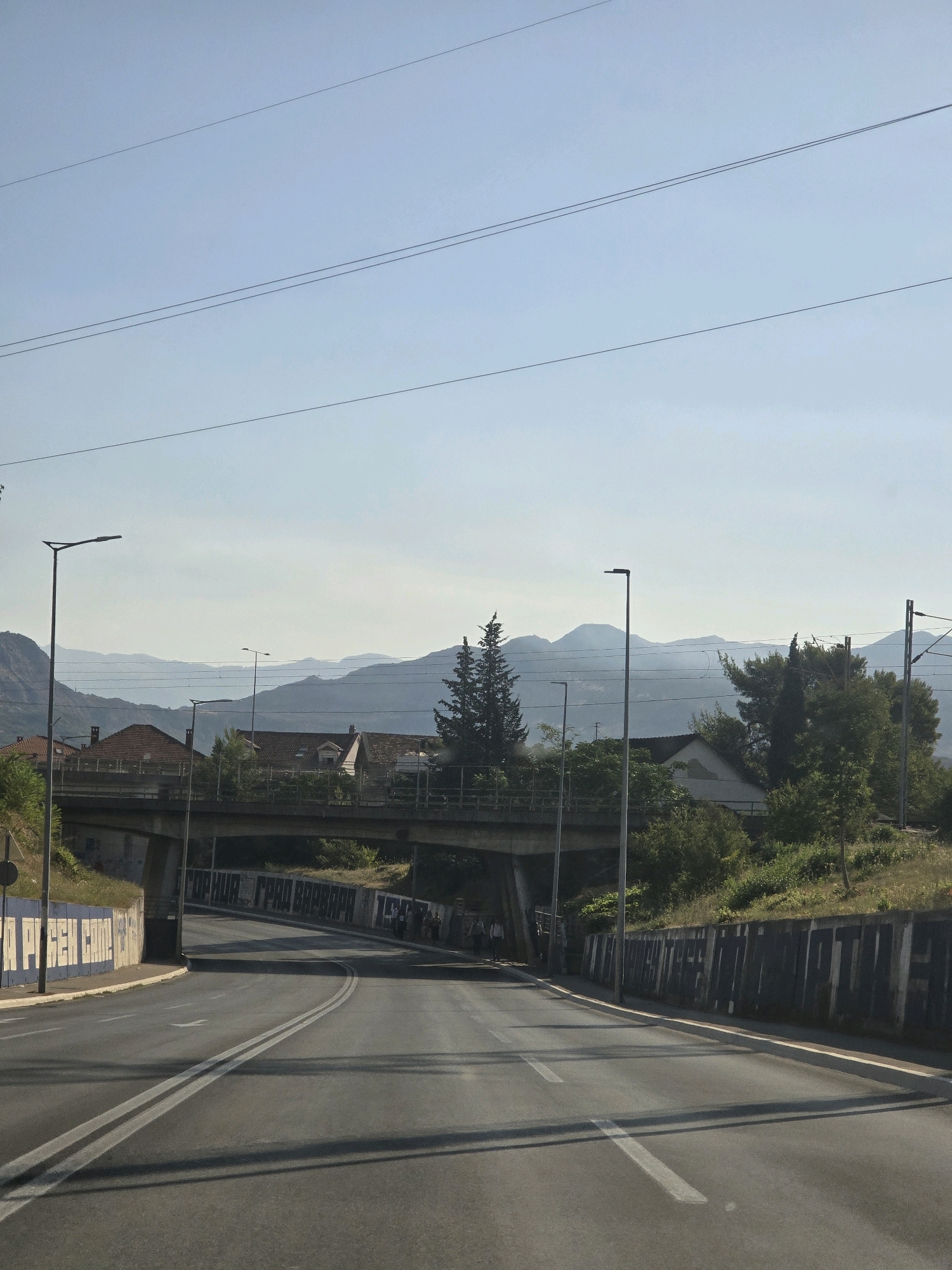The image depicts a road in Podgorica, Montenegro, with a clear blue sky. In the foreground, the road curves to the right. On the left side of the road, there is a concrete wall with blue and white graffiti. Further down the road, a concrete overpass crosses overhead. On the right side of the road, there is a retaining wall also covered in graffiti. Beyond the overpass and retaining wall, there are houses with red-tiled roofs, trees, and lush green vegetation. In the background, layers of mountains are visible, fading into the hazy sky. Several streetlights are positioned along the road and near the overpass. Power lines are strung across the sky. A few people can be seen near the retaining wall on the right side of the road.