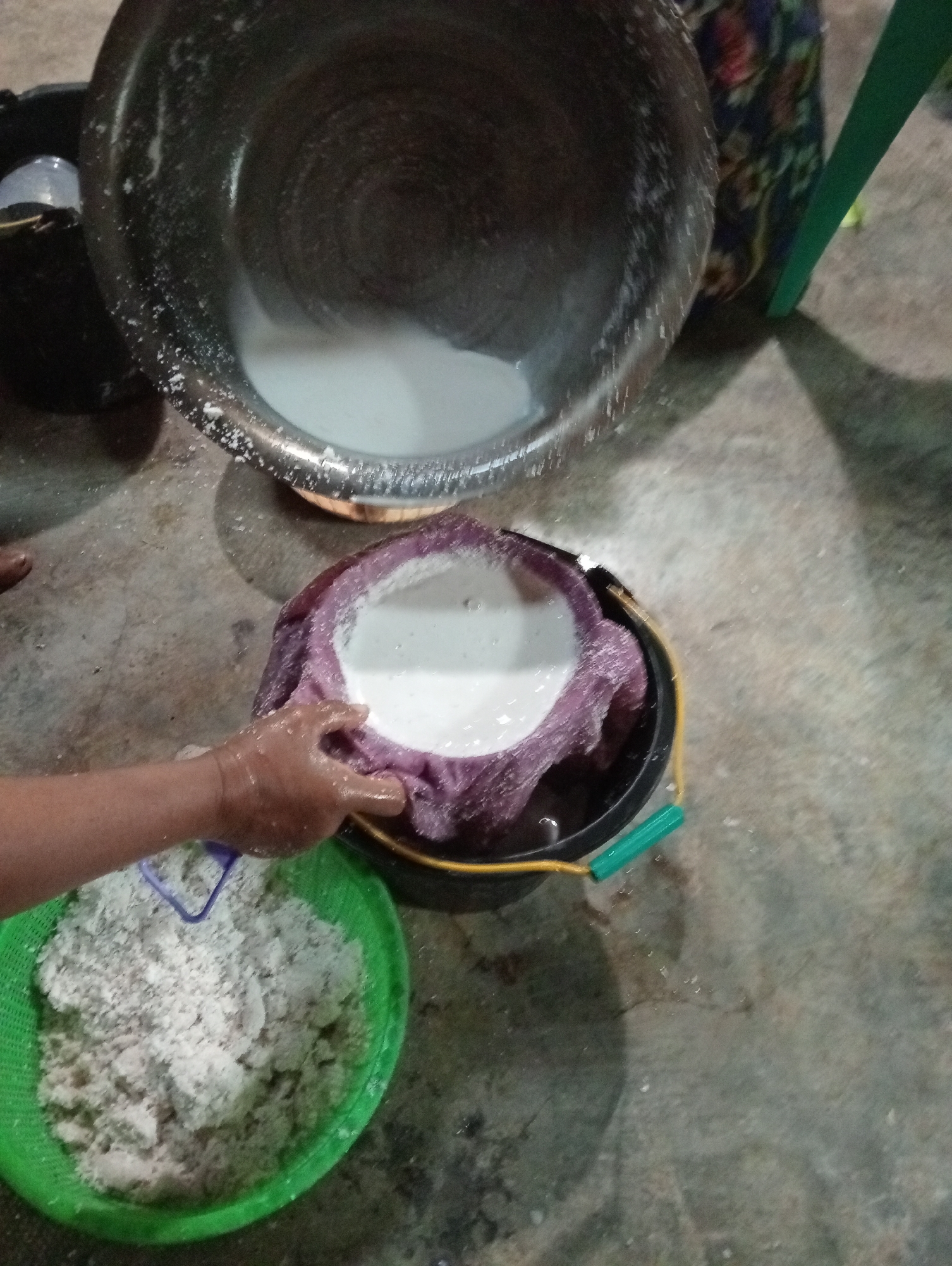 In Campago Barat, Indonesia, a person's hand is seen squeezing a purple cloth filled with a milky liquid. The cloth is placed over a black bucket with a green handle, and the liquid is straining into the bucket. To the left, a green basket contains a pile of grated coconut. In the background, a large metal bowl holds more of the milky liquid, with some of the liquid and grated coconut spilled onto the concrete floor. The scene appears to be an outdoor setting, possibly a kitchen or preparation area, given the concrete ground and simple containers.