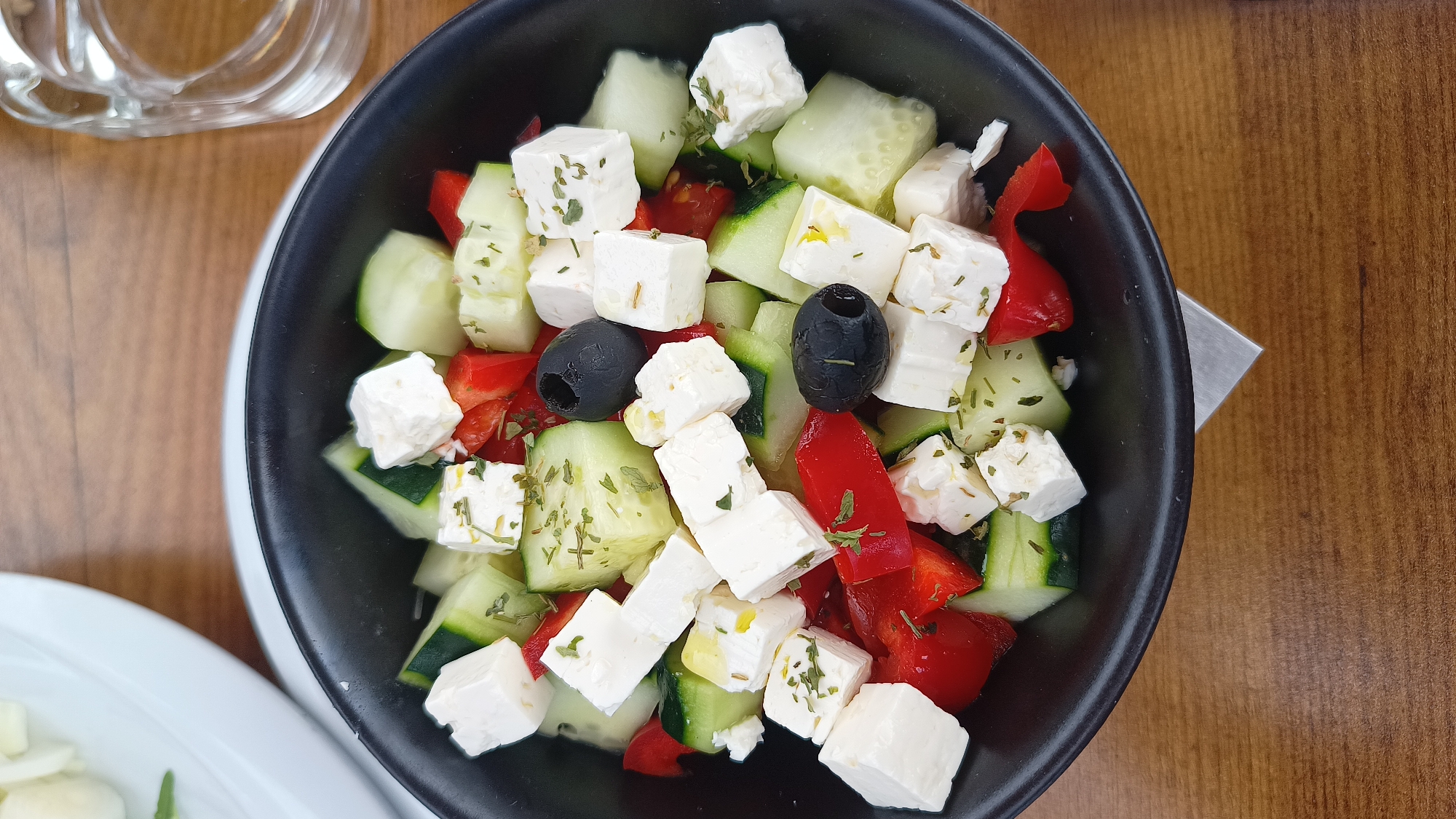 A vibrant, top-down close-up shot captures a fresh salad served in a black bowl, resting on a warm wooden table. The salad, characteristic of a Greek-style dish, features a generous assortment of cubed white feta cheese, chopped green cucumbers, and bright red bell pepper pieces. Several glossy black olives are interspersed among the colorful ingredients. Many of the feta cubes and cucumber slices are sprinkled with dried green herbs, likely oregano, and a light, oily dressing is visible, enhancing their appeal.
In the upper left background, part of a clear glass, possibly for water, is visible, reflecting ambient light. To the lower left, a portion of a white plate with additional food items can be seen, suggesting a larger meal. A small metallic object, possibly a utensil or part of a table accessory, peeks out from under the black bowl on the right side. The wooden table surface provides a natural and inviting backdrop to the colorful dish. The image presents the salad in a well-lit environment, ready for consumption, likely in a dining establishment in Umag, Croatia. No people or visible text are present in the image.