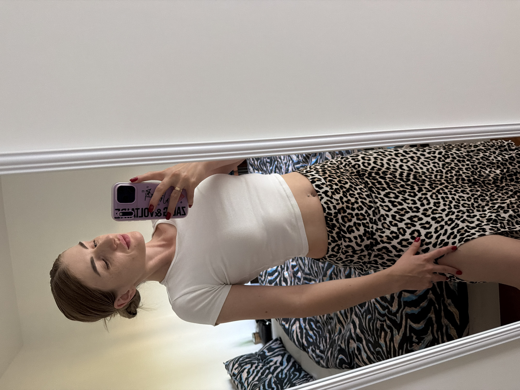 A woman stands in front of a mirror, taking a selfie. She is wearing a white crop top and a leopard-print skirt with a high slit. Her hair is pulled back in a bun, and she is wearing red nail polish. The background shows a bedroom with a bed covered in a patterned comforter and a wall with a mirror.