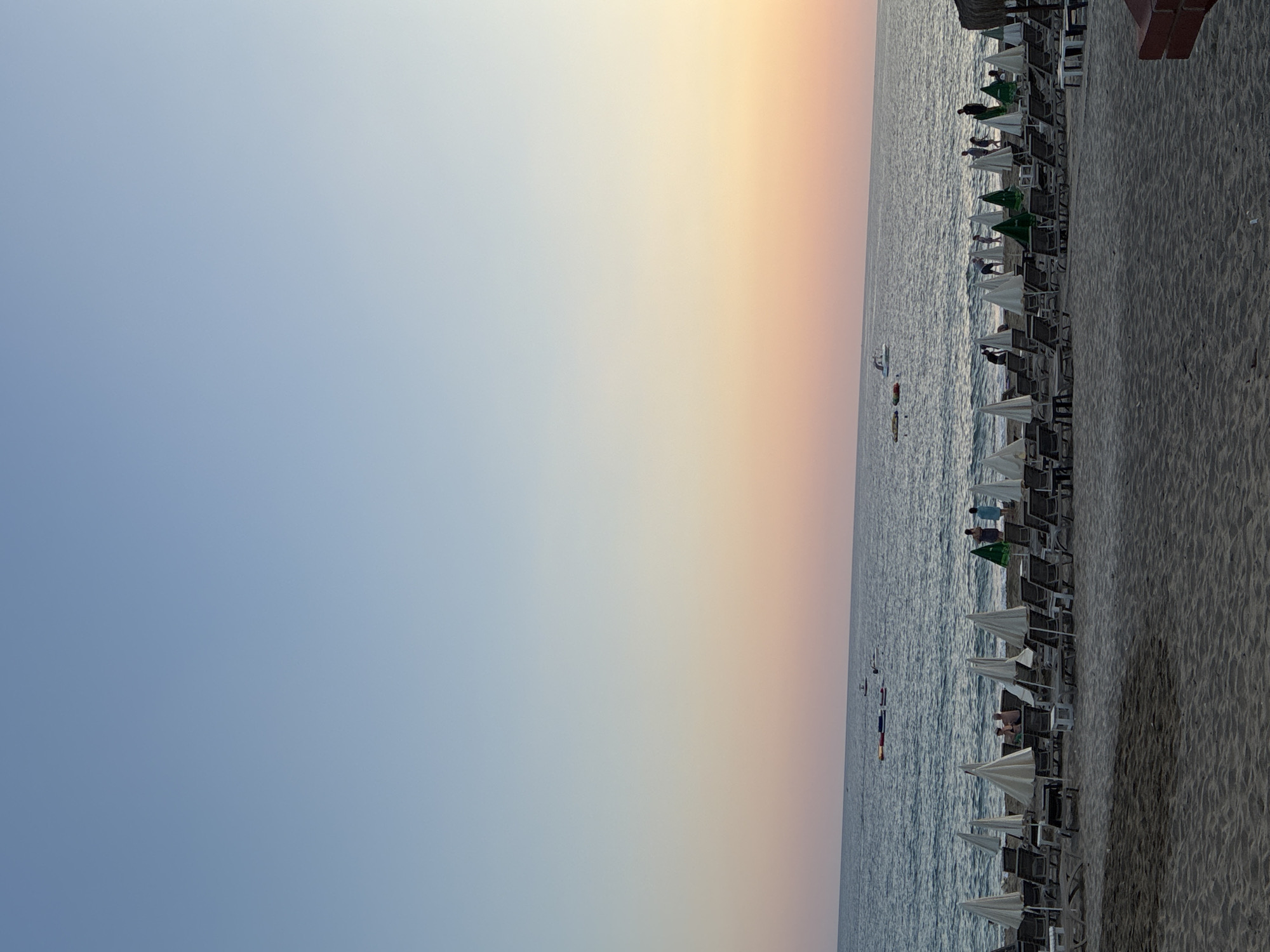 The image depicts a wide sandy beach in Alanya, Turkey, with rows of empty lounge chairs and closed umbrellas stretching towards the sea. The sea is calm, with a few small boats visible in the distance. The sky transitions from a clear blue at the top to a soft orange and pink gradient near the horizon, indicating either sunrise or sunset. Several people are scattered across the beach and in the shallow water, some appearing to be engaging in recreational activities. The overall atmosphere is peaceful and serene, with the natural beauty of the coastline taking center stage.