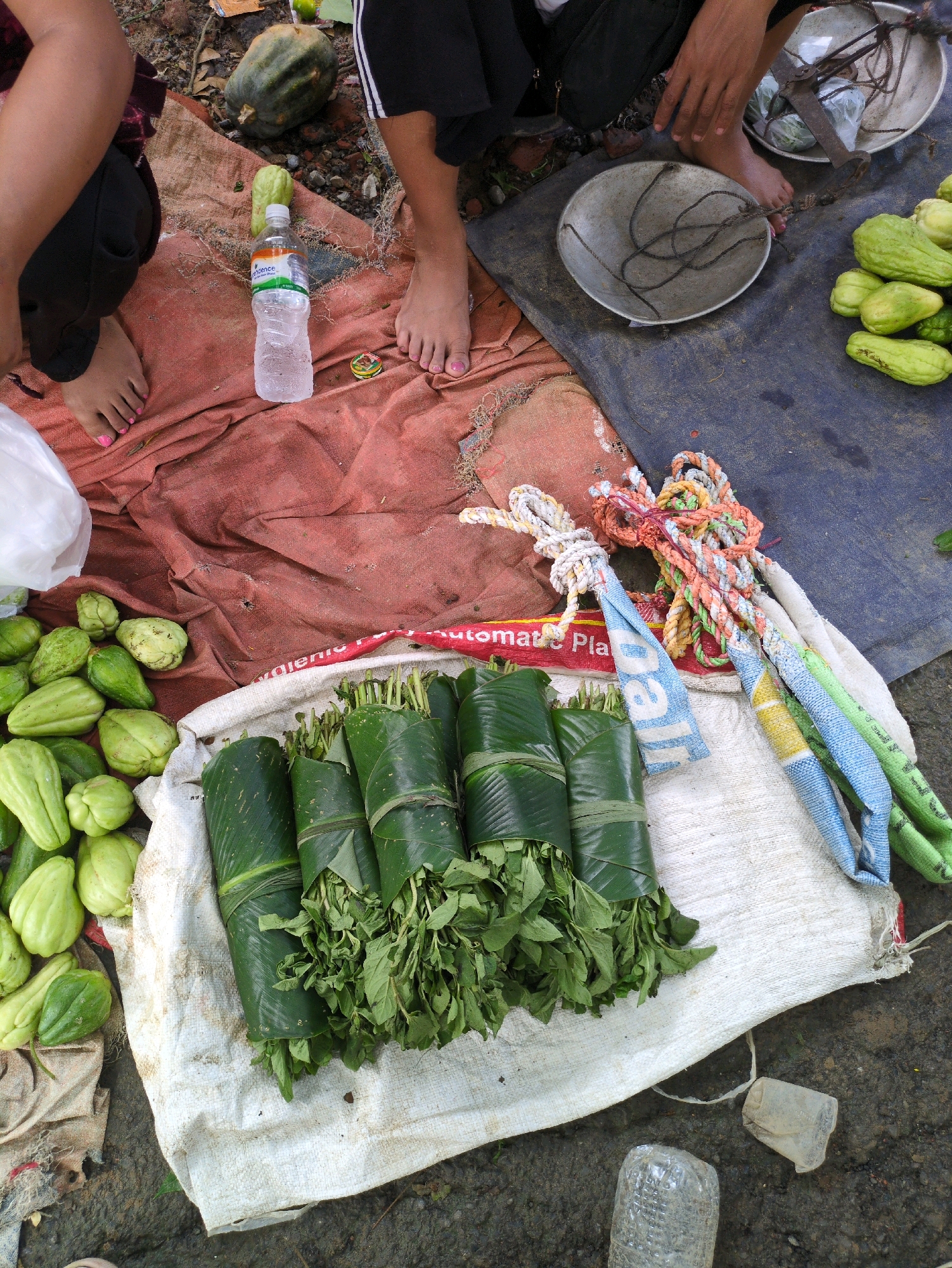 In Borchora, India, a market stall displays produce and goods on the ground. A person's legs, one with painted toenails, are visible on a red cloth, suggesting they are sitting or kneeling. Various vegetables, primarily green and elongated like chayote squash, are arranged on the ground, some wrapped in banana leaves. Several bundles of leafy greens are also present, some tied with string. A plastic water bottle, a scale with a metal bowl and ropes, and colorful braided ropes are among the items scattered around. The ground is a mix of dirt, gravel, and a dark blue mat. The overall impression is a casual market setting with fresh produce for sale.