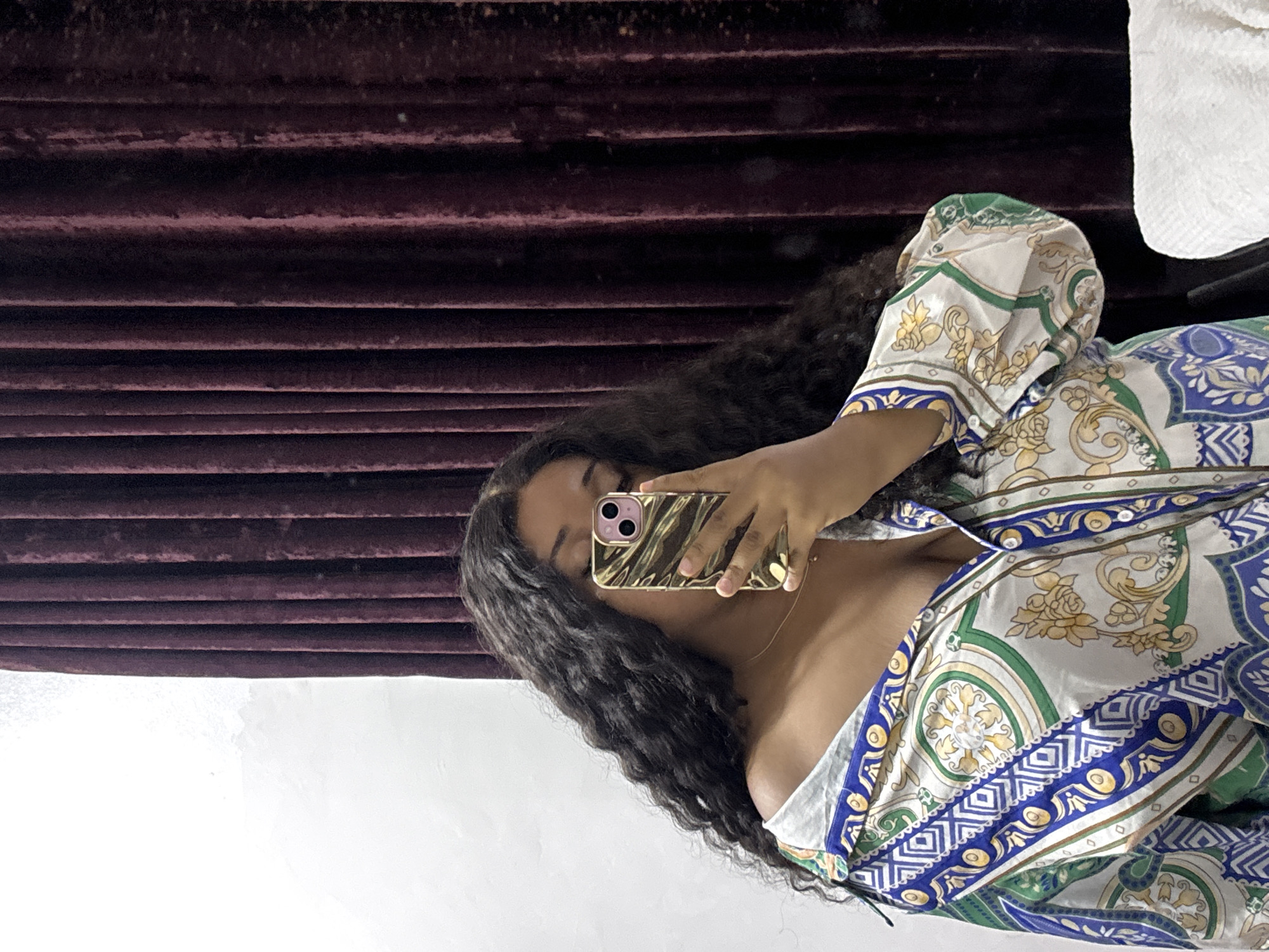 A woman with long, dark, curly hair is taking a selfie with her iPhone, partially obscuring her face. She is wearing a white, blue, and green patterned blouse. The background consists of a dark burgundy textured wall. The location in the city of Port Harcourt, Nigeria cannot be determined from the image.