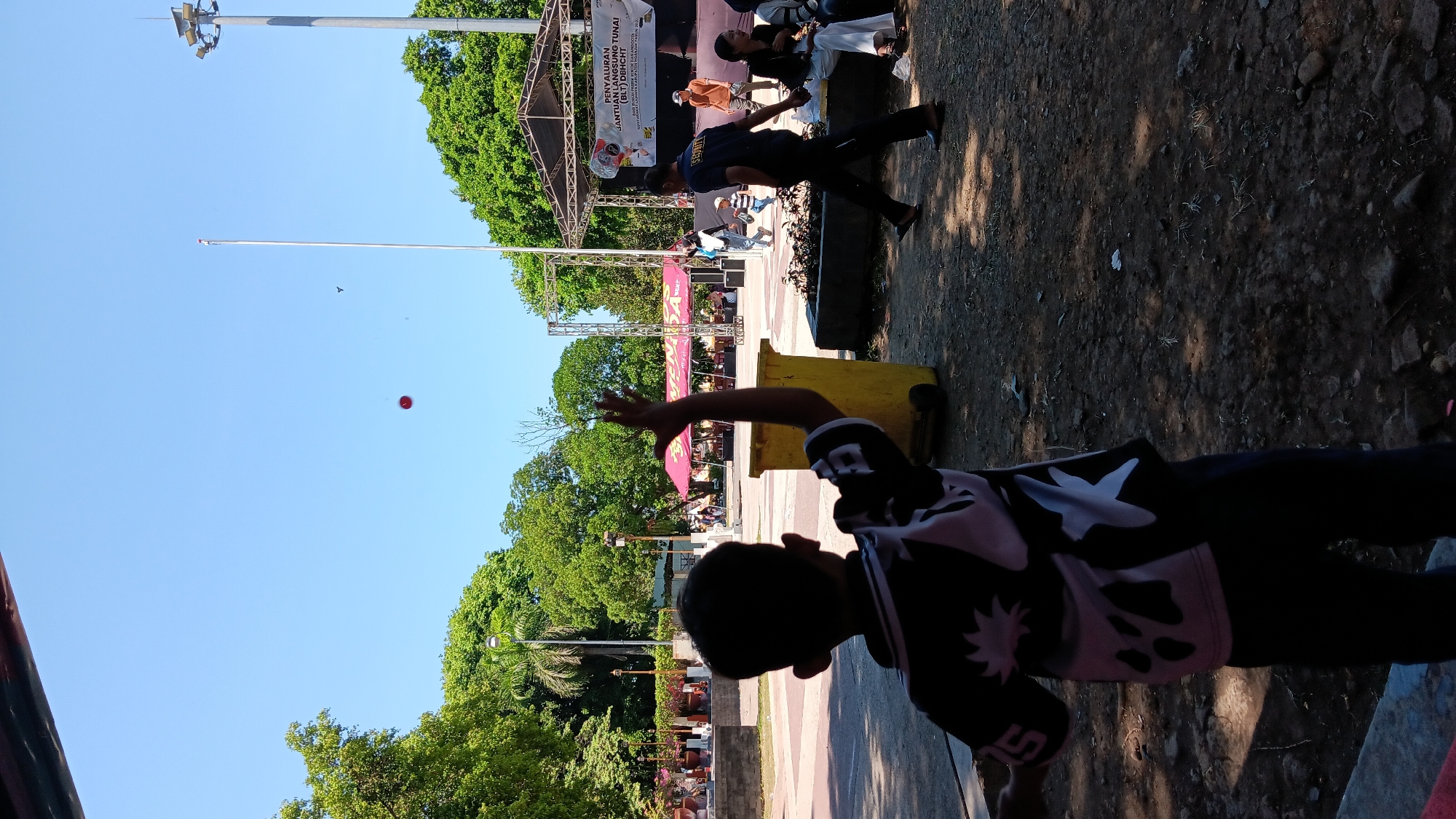 The image shows an outdoor scene, likely in a park or public square in Nganjuk, Indonesia. In the foreground, a young boy is silhouetted, facing away from the camera and looking upwards. He is wearing a dark t-shirt with a white, abstract pattern and dark pants. His right arm is raised, as if he is throwing something or waving.
Above the boy, a small red ball is visible in the clear blue sky. The background features a stage set up with a banner that reads "PENYALURAN BANTUAN LANGSUNG TUNAI (BLT DN)...". Several people are gathered on and around the stage, including individuals who appear to be staff or performers, and others sitting in the audience. A security guard in a dark uniform is walking across the scene.
To the left, there are trees, a lamppost, and some decorative planters. A large yellow trash bin is positioned near the boy. The ground is a mix of dirt, gravel, and scattered debris, with some areas covered in shade from overhead structures. The overall atmosphere suggests a public event or gathering taking place on a sunny day.