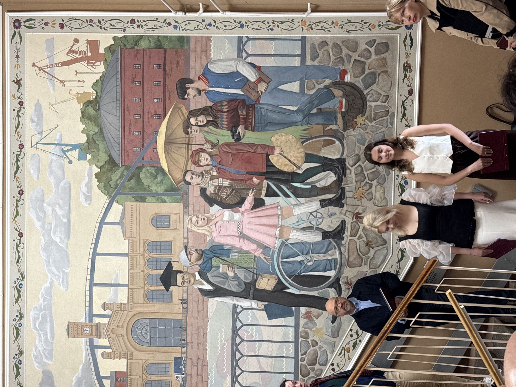 The image depicts a large mural on a wall, likely in a public building, possibly a museum or a university in Saint Petersburg, Russia, given the style and imagery. The mural is divided into two main sections. The upper section features a cityscape with a prominent clock tower, industrial cranes, a red brick building with a domed roof, and a bridge spanning a body of water. The lower section of the mural shows a group of people in casual attire, some holding items like a bicycle, a palette, a camera, and books. The people are depicted in a narrative scene, perhaps representing students or a diverse group of individuals engaged in various activities.
In the foreground of the photograph, superimposed over the lower part of the mural, are several people standing on a staircase. These individuals are dressed in contemporary clothing. A man in a dark jacket and glasses is holding a book. To his right, a woman with blonde hair is wearing a black top with voluminous white sleeves and a white skirt, also holding a book. Further to the right, another woman with dark hair is wearing a white vest over a dark skirt and holding a book. A fourth woman, with blonde hair and wearing a beige coat, is also visible. The presence of these individuals and the staircase suggests the photograph was taken in a physical space where the mural is displayed, rather than just a representation of the mural itself.